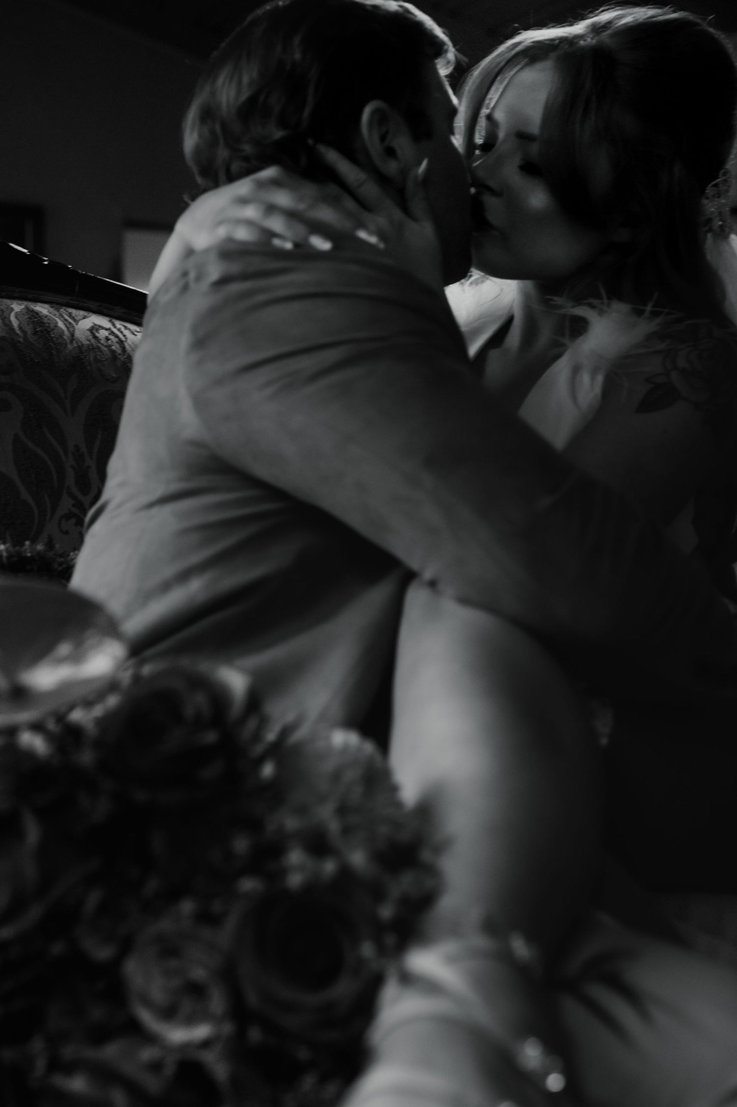 Intimate black and white wedding photo showing a romantic kiss with a bouquet visible in the foreground.