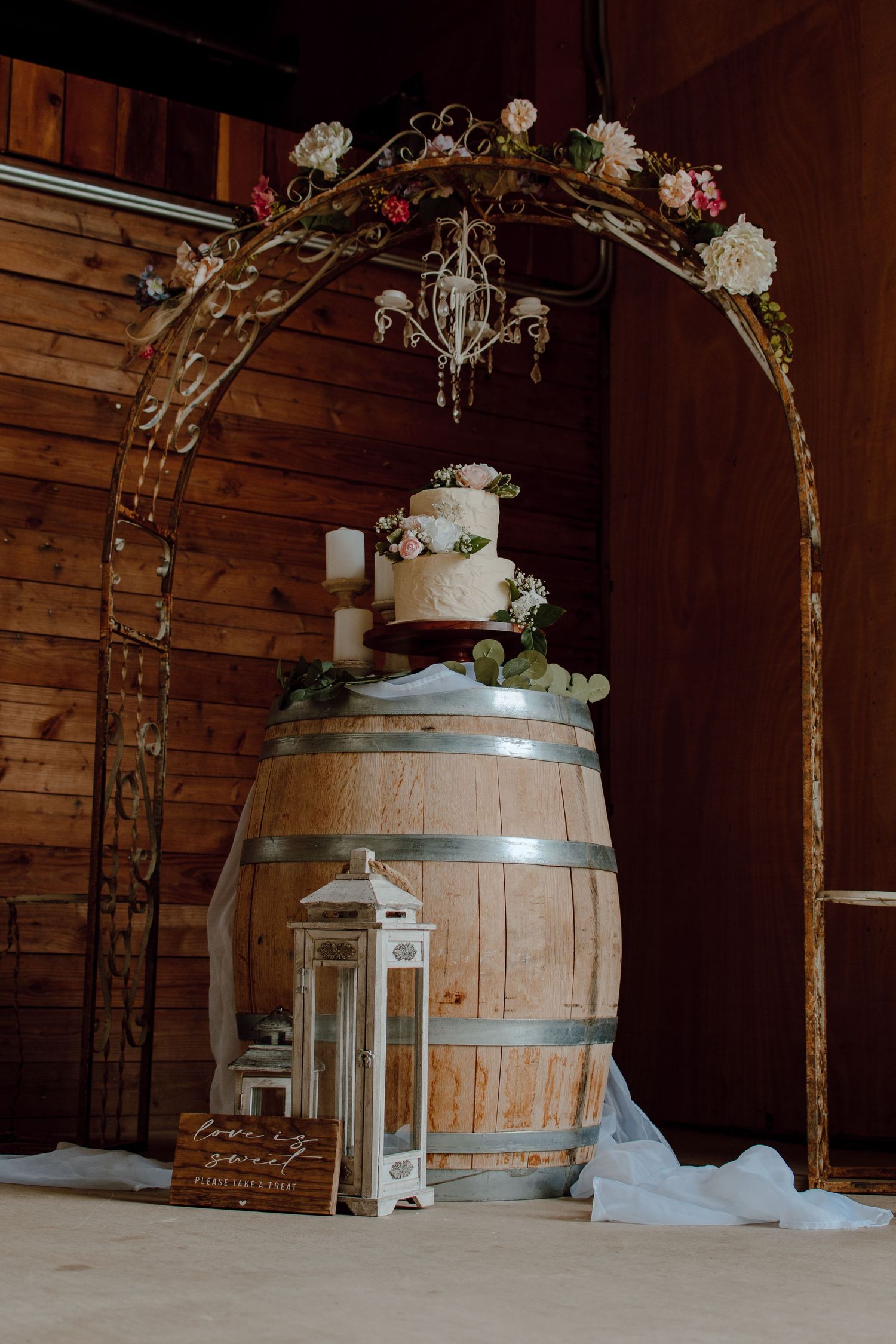 Wedding cake displayed on wooden barrel under floral arch with lantern decor.