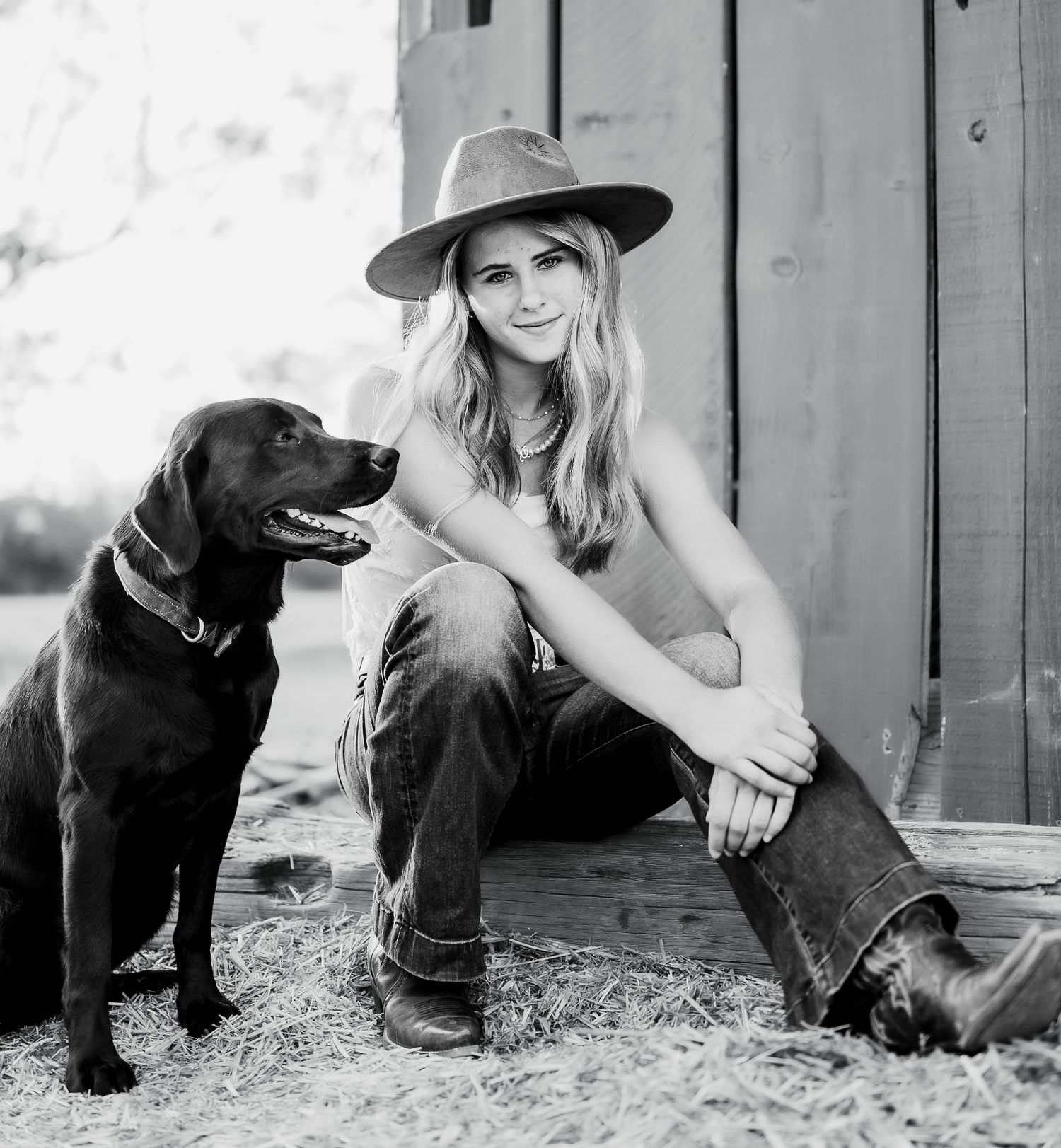 Black and white photo of a person wearing a hat sitting next to a black labrador retriever dog on rustic wooden steps.