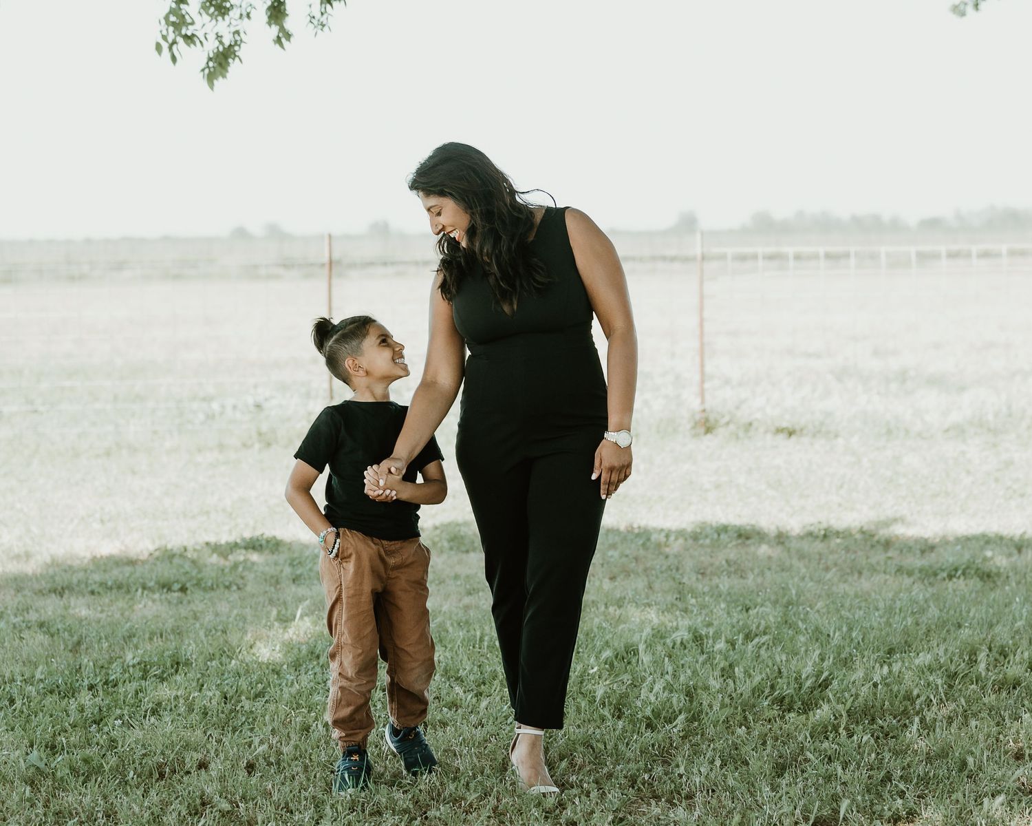 Parent and toddler in matching black outfits walk together on grassy hill during outdoor photo session.