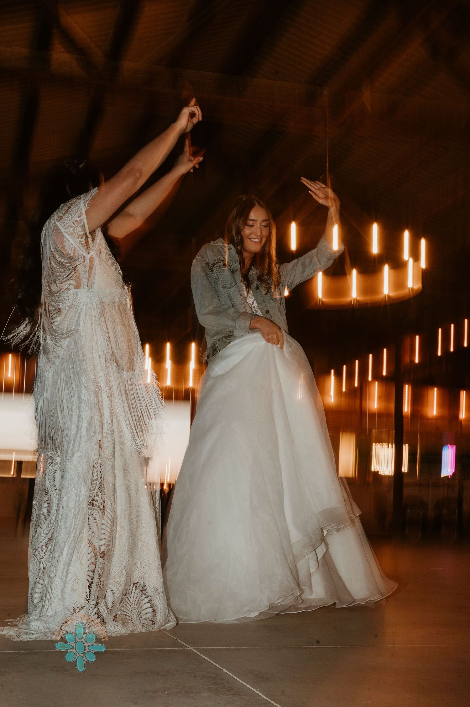 Two people in white wedding dresses dancing together under warm candlelight in a rustic venue.