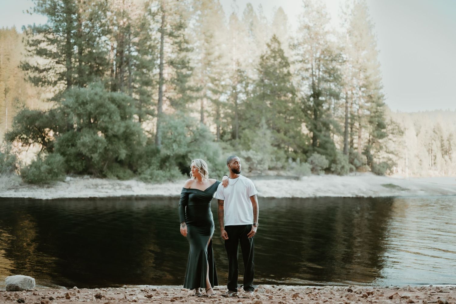 A couple stands together on a snowy lakeshore with evergreen trees in the background during winter.