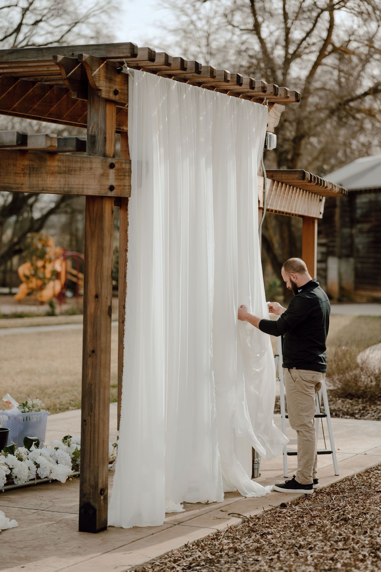 Setting up white drapes on a wooden pergola for an outdoor event.