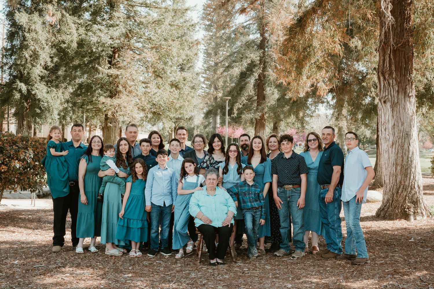 A large group wearing coordinated teal and blue outfits poses together outdoors among tall trees in a forest setting.