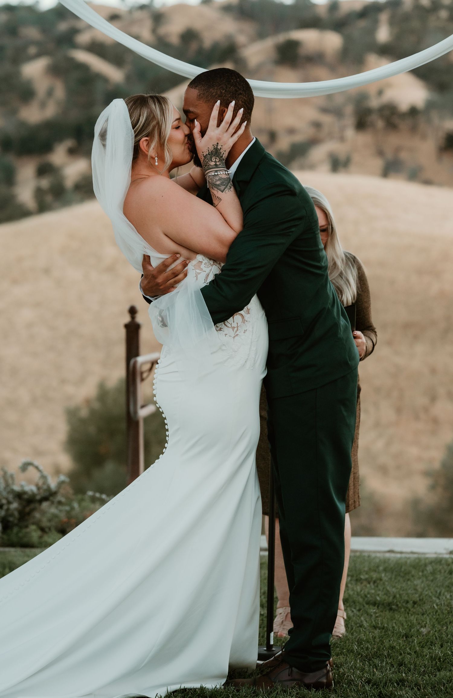Romantic outdoor wedding ceremony with couple kissing under a white arch against rolling hills.