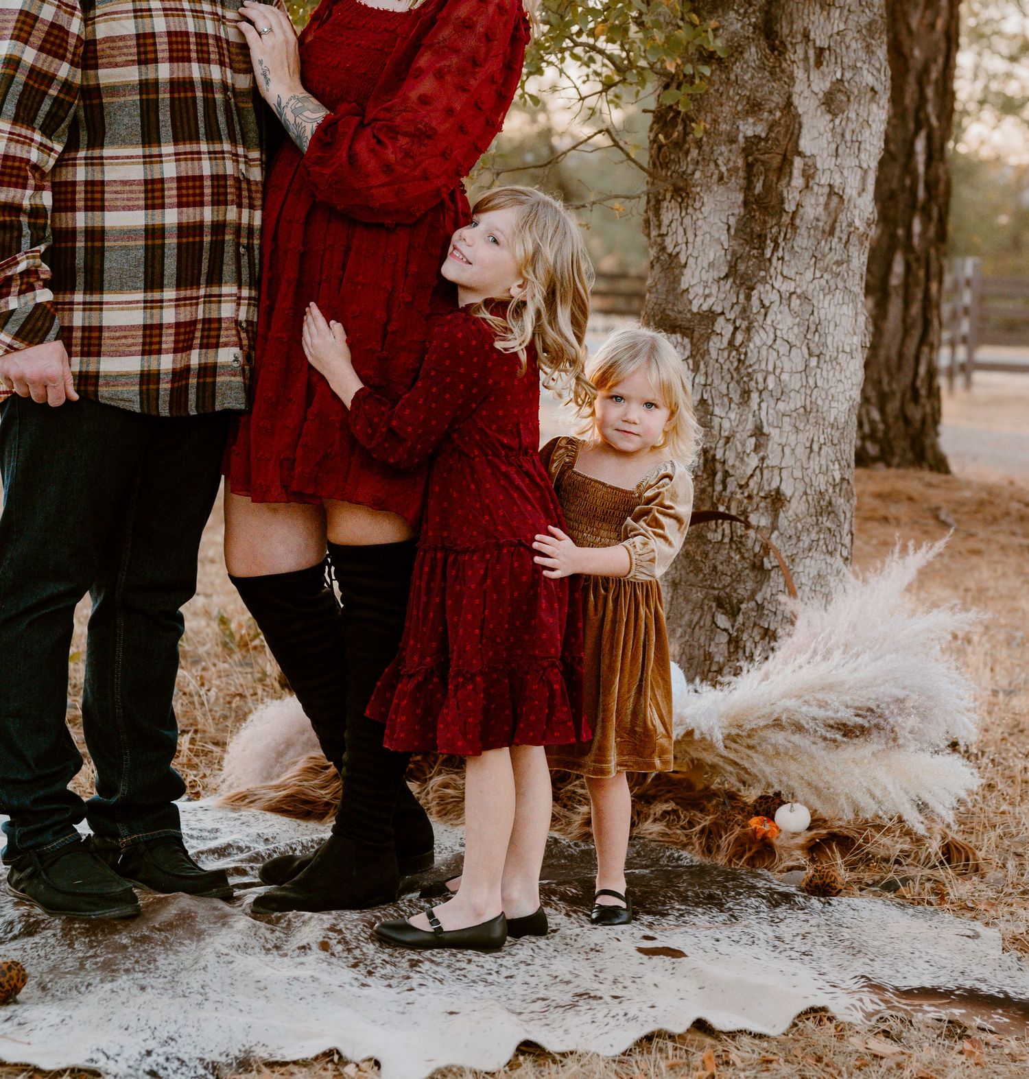 A family wearing red and plaid outfits stands together near a tree trunk in an outdoor autumn setting.