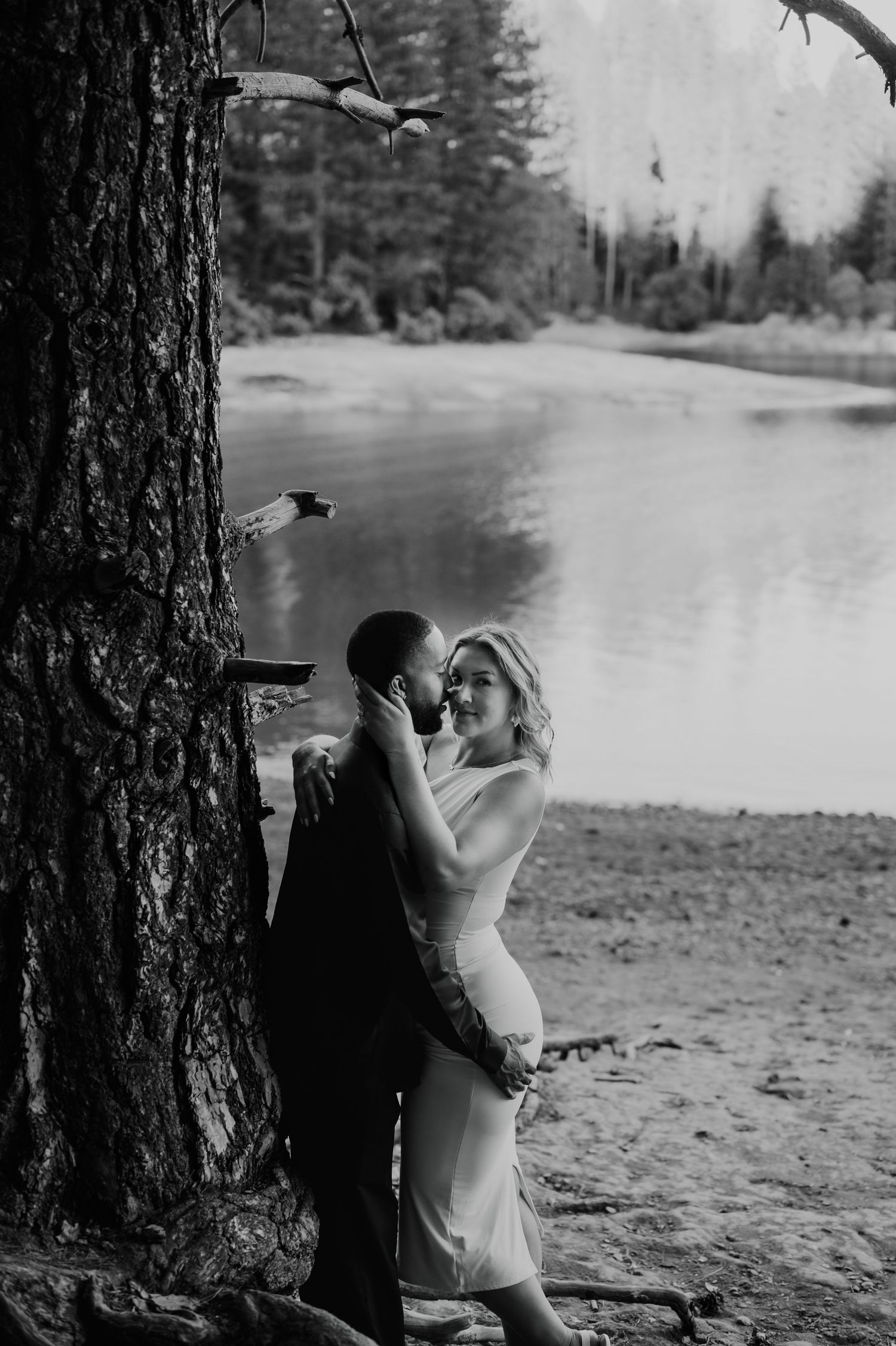 A romantic black and white moment captured by a lake with trees and nature surrounding the couple.