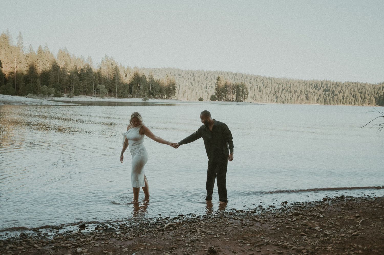 Two people holding hands walk along a tranquil lakeside at dusk with evergreen trees in the background.
