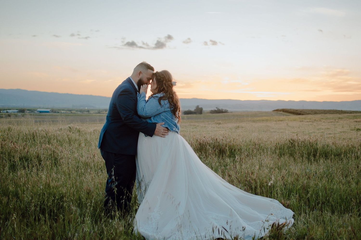 A romantic sunset wedding photo in a grassy meadow with mountains in the background and a flowing white dress.