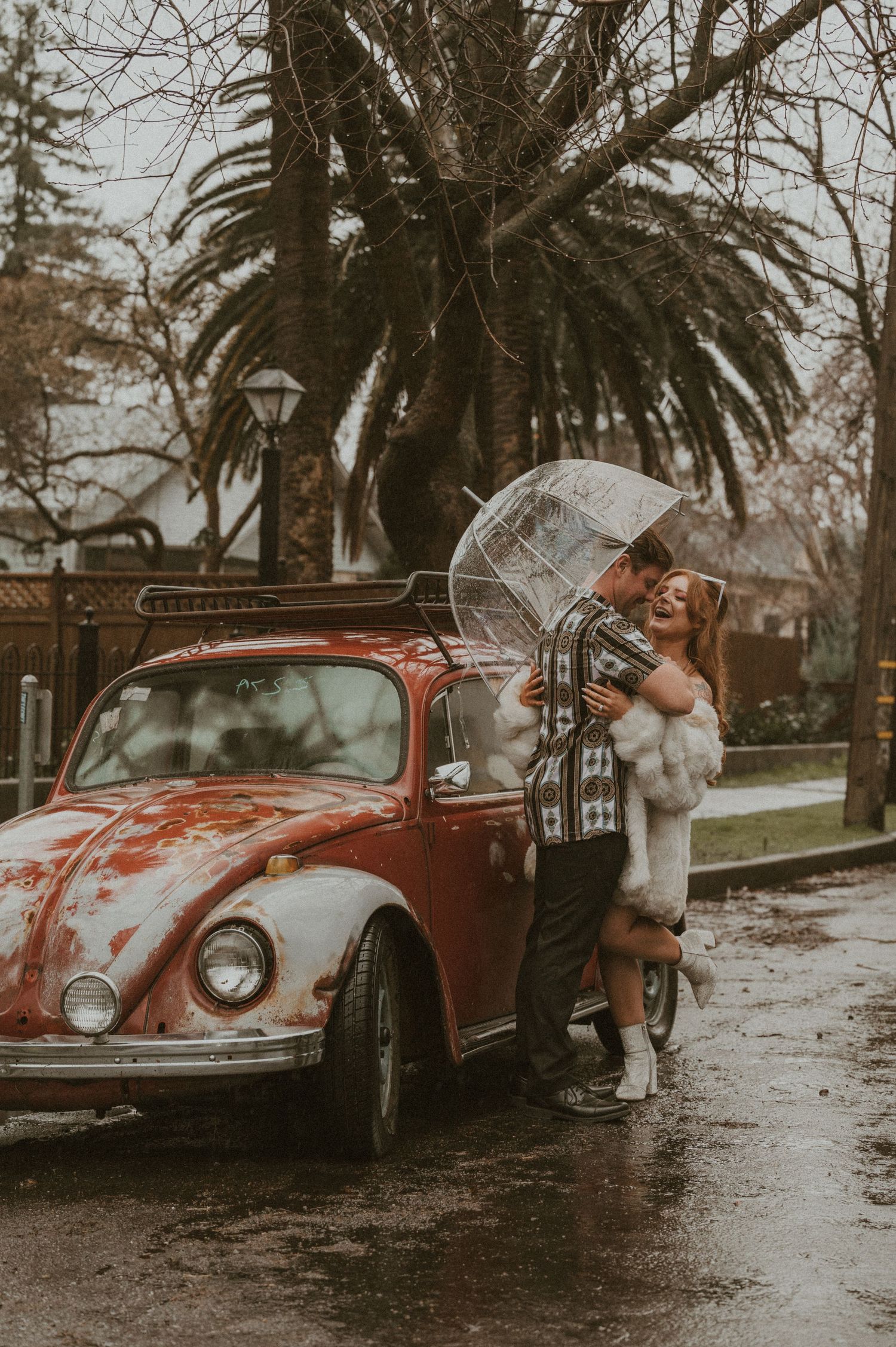 A vintage Volkswagen Beetle and person photographed in a moody rainy setting with palm trees in the background.