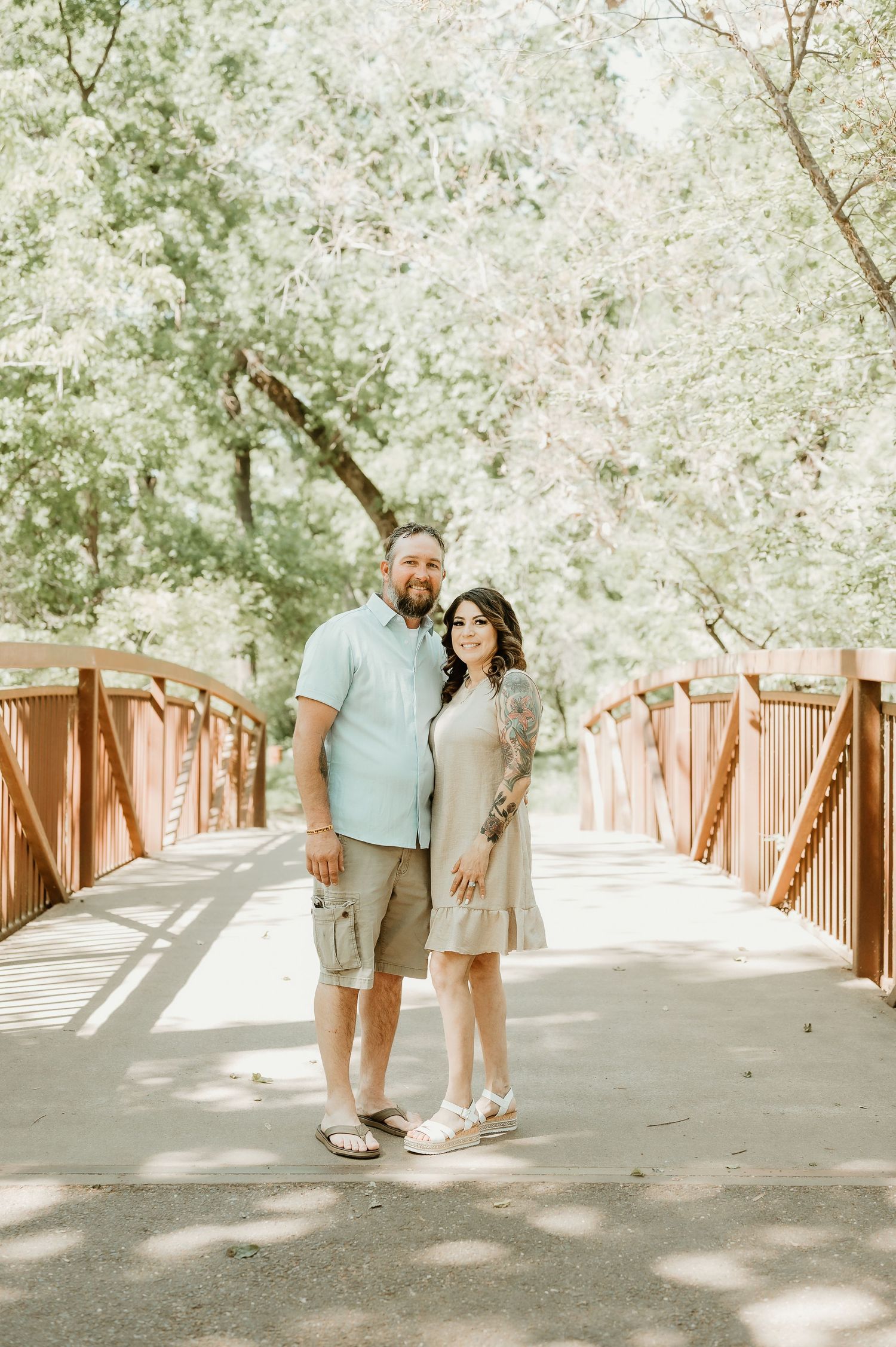 Couple poses along a wooden bridge path surrounded by blooming white spring trees on a sunny day.