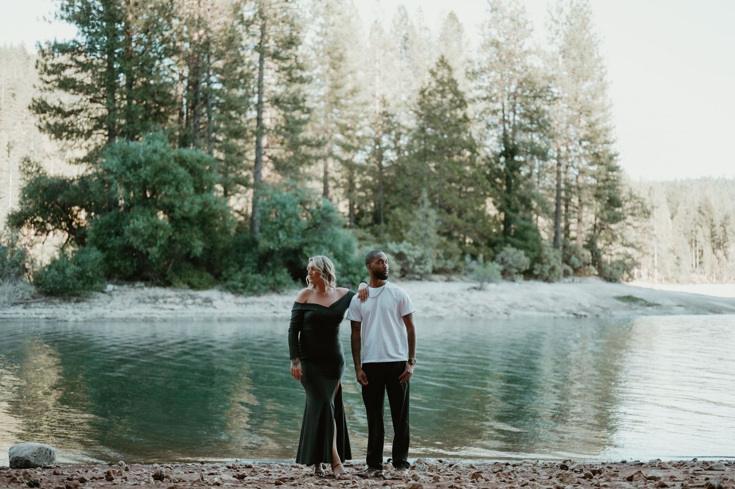 A group of people stroll along a picturesque lakeside path with evergreen trees and mountains in the background.