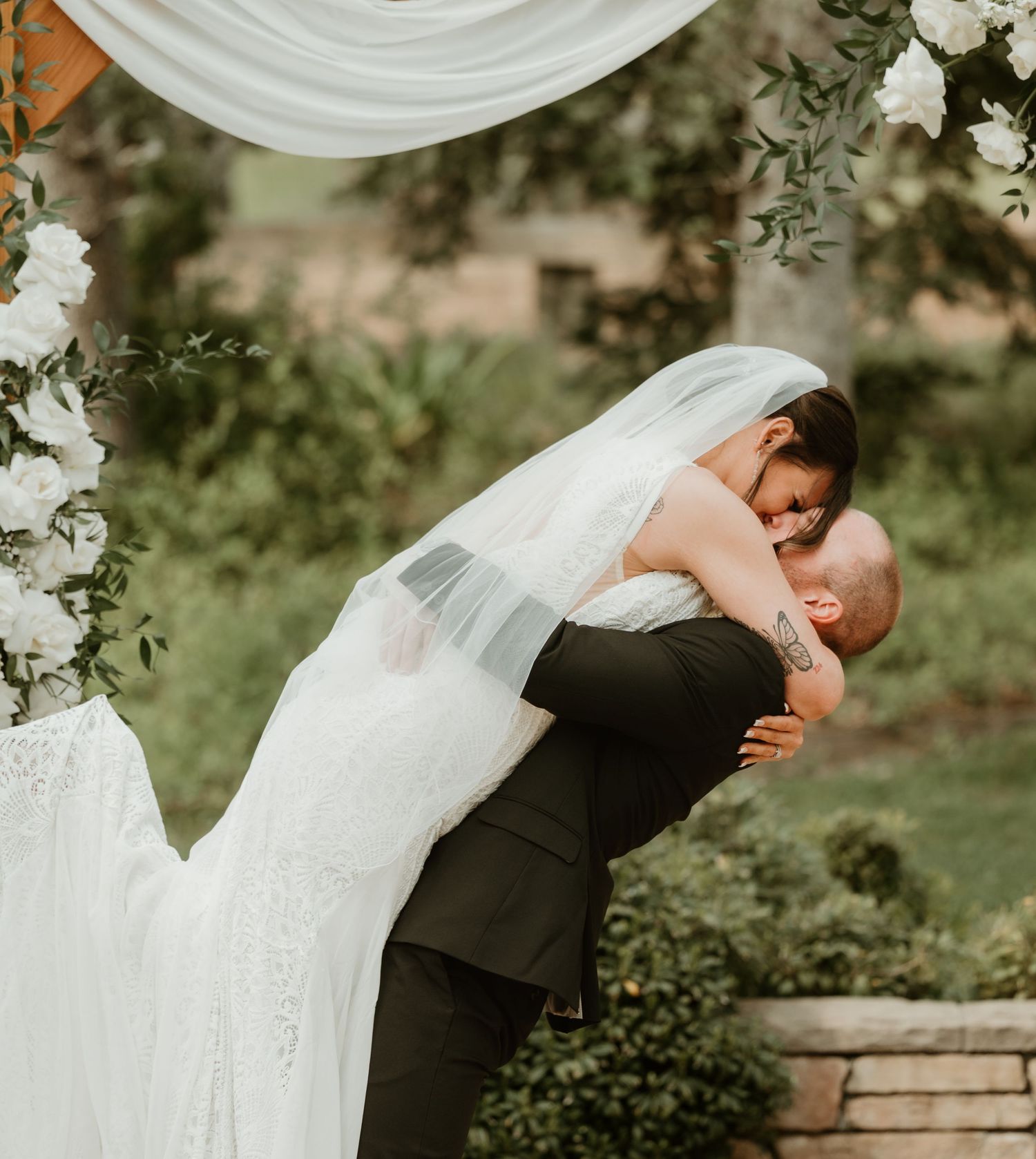 A bride and groom share a romantic dip pose in a garden setting with flowing white veil and formal attire.