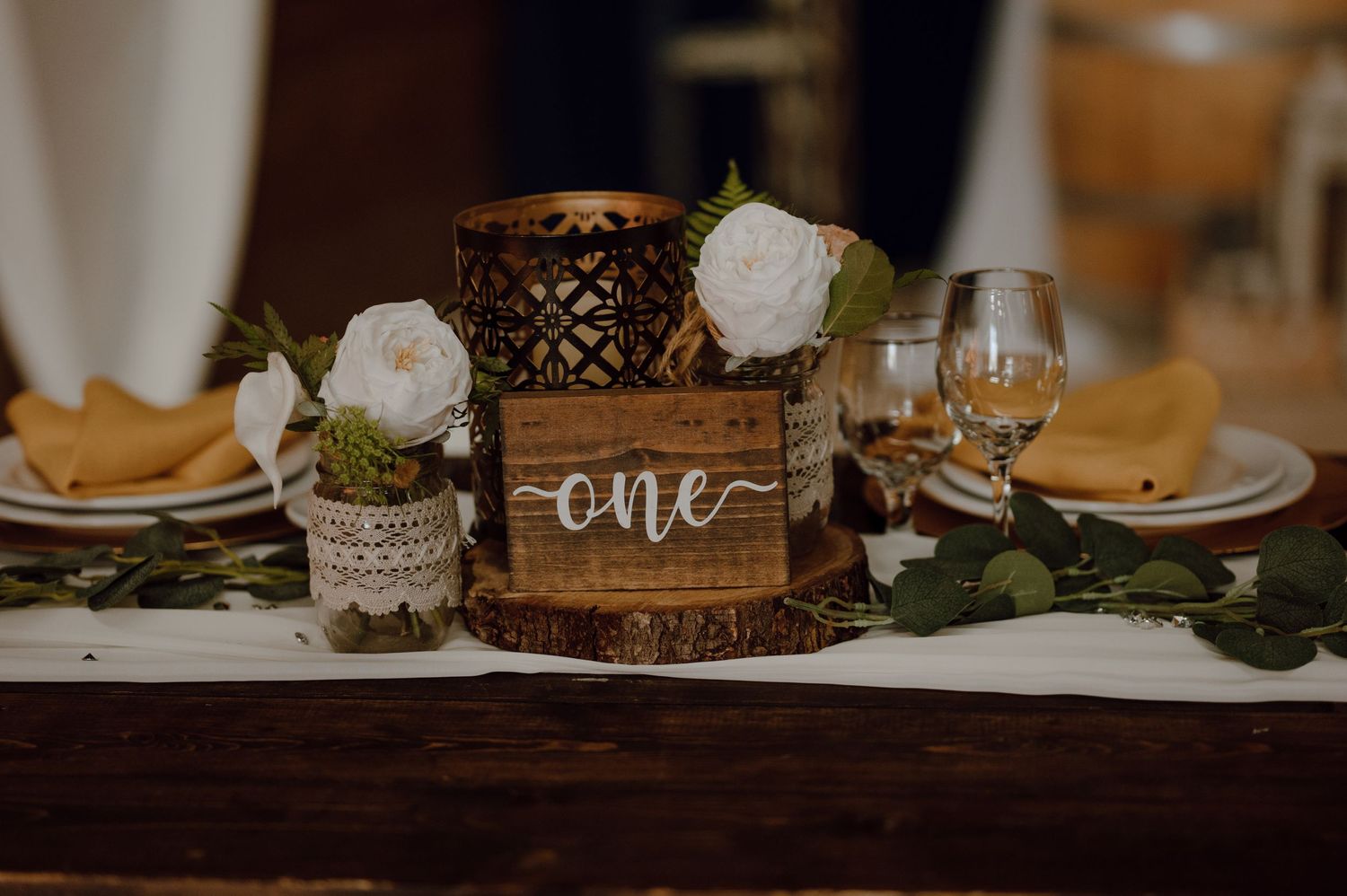 Rustic wedding table setting with wooden 'one' sign, white roses in mason jars, and vintage lantern centerpiece.