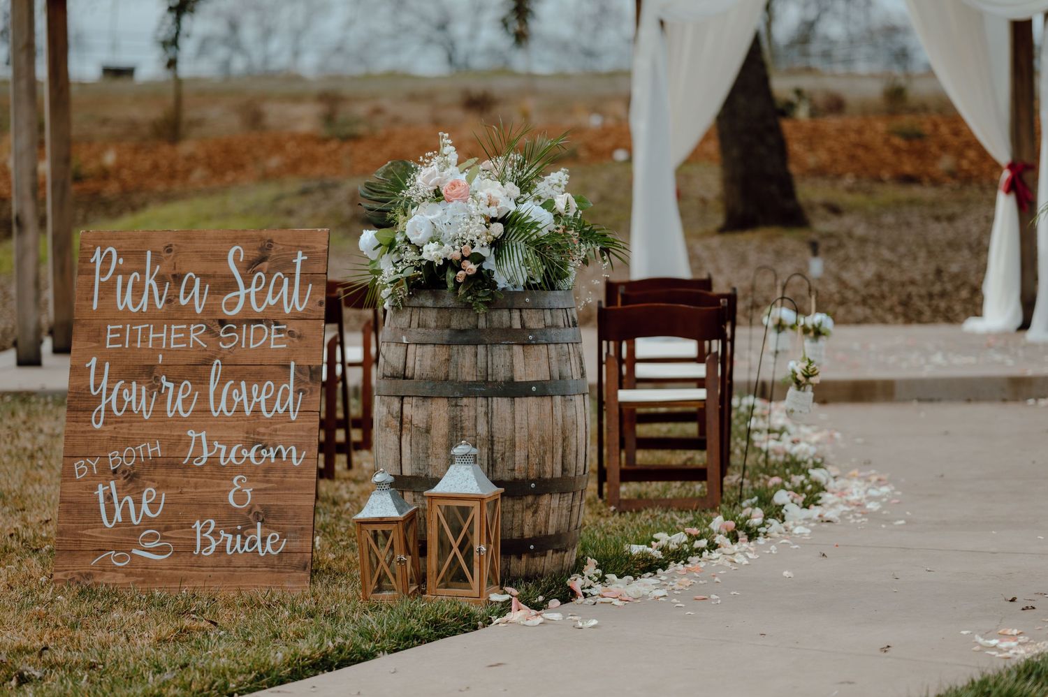 Wooden welcome sign with lantern and floral decorations at an outdoor wedding ceremony entrance.
