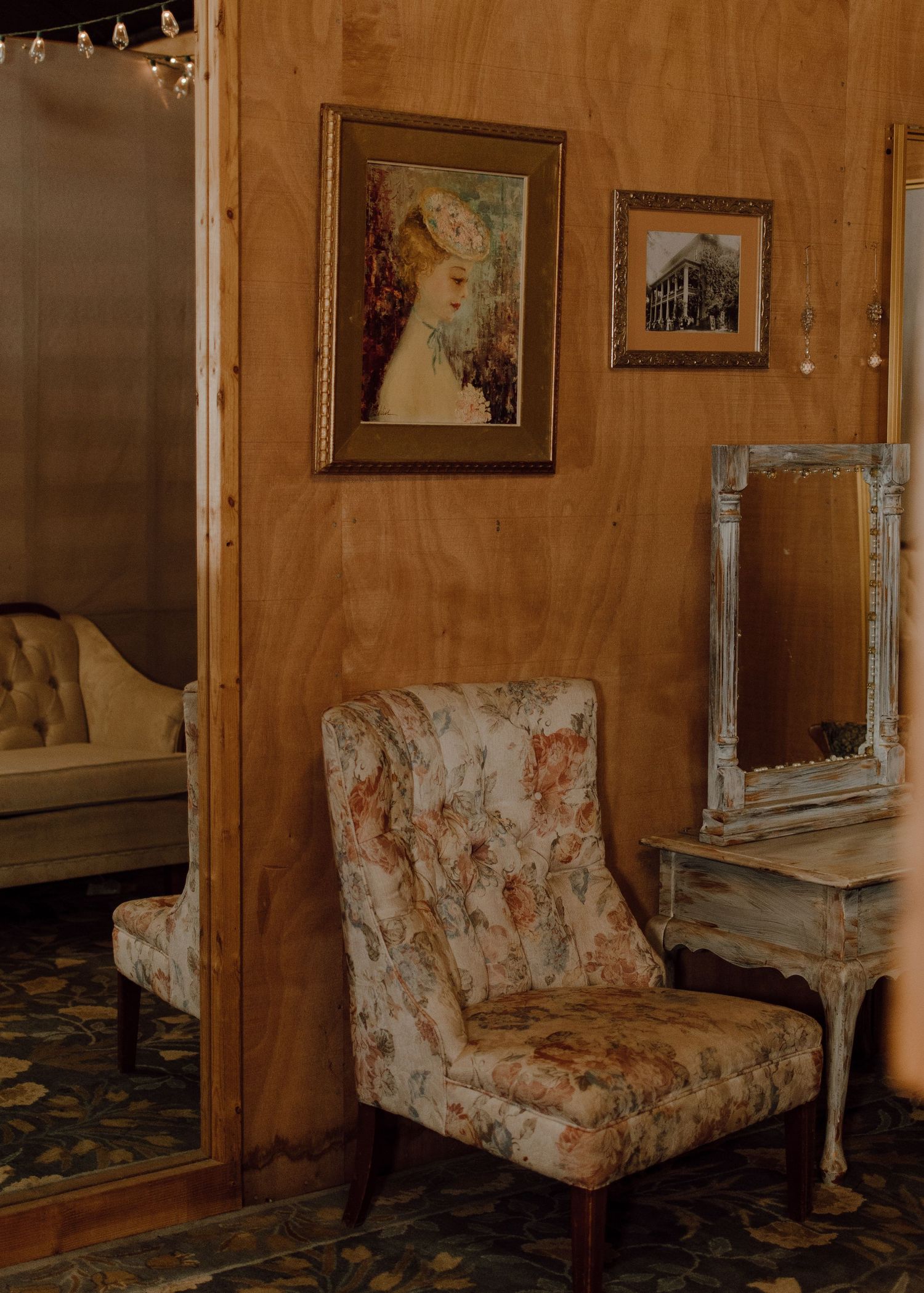 Vintage-style sitting area with floral upholstered chair, framed artwork, and distressed mirror against wood paneling.
