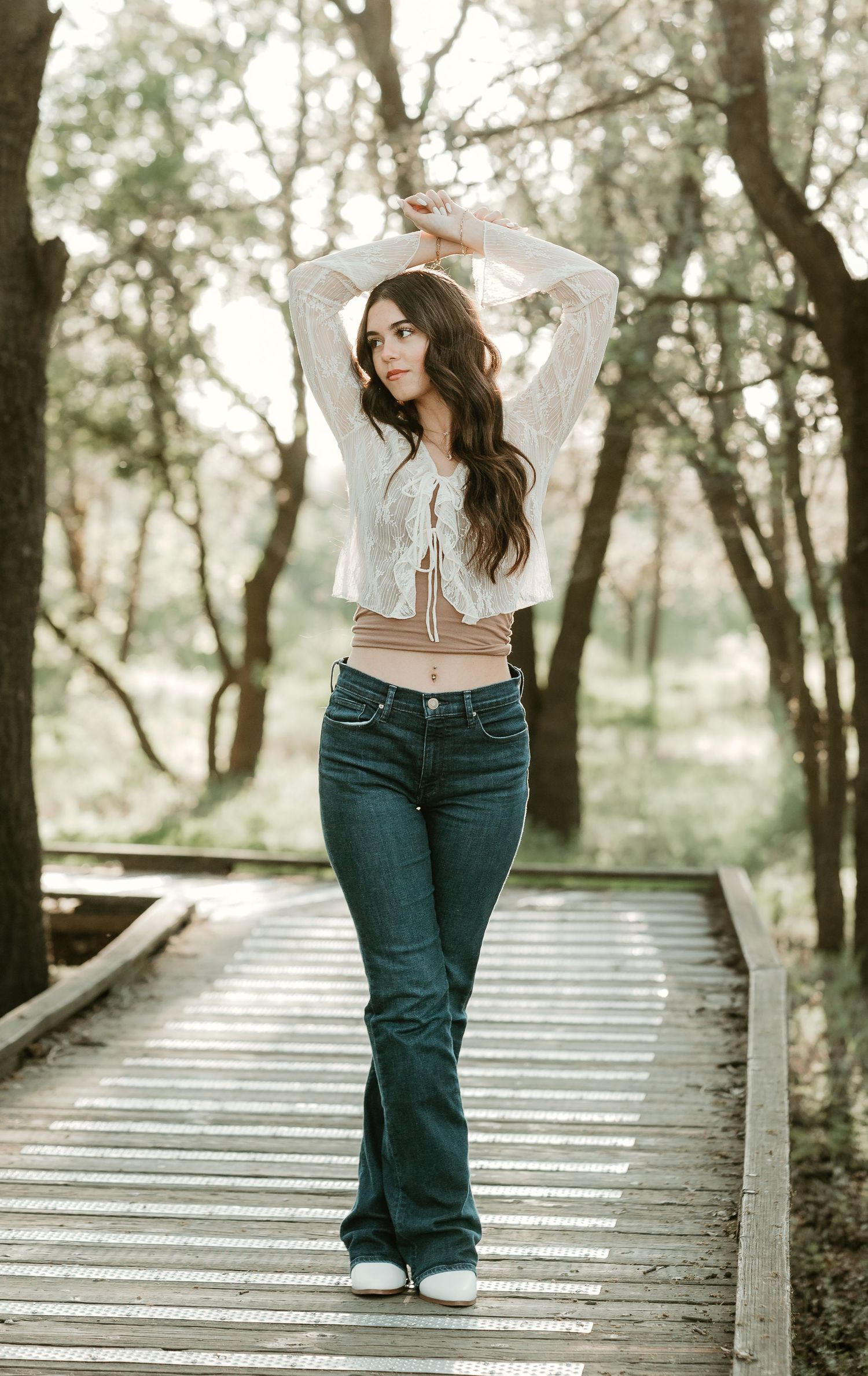 Young woman in white lace crop top and flare jeans posing on wooden boardwalk surrounded by trees.