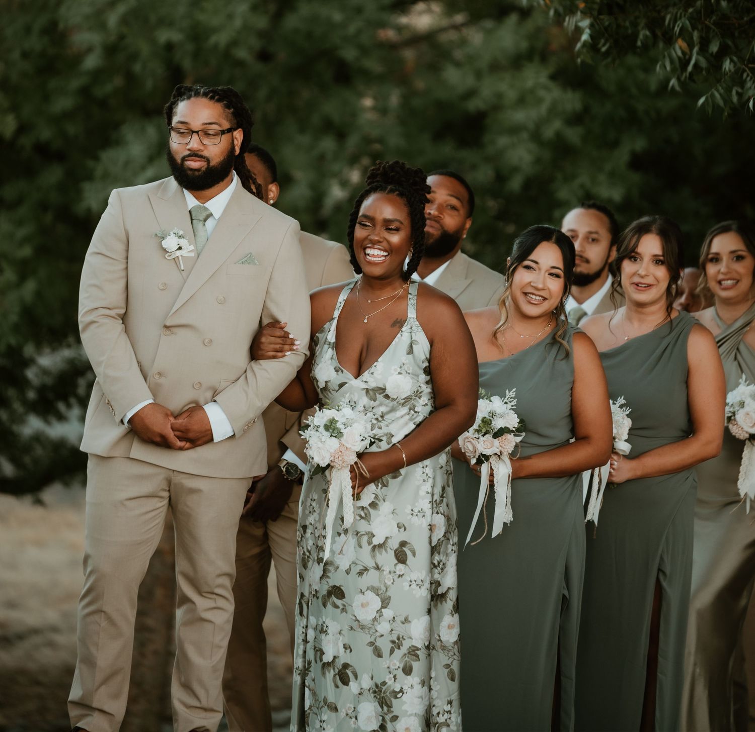 Wedding party in sage green dresses and tan suits posing together outdoors.