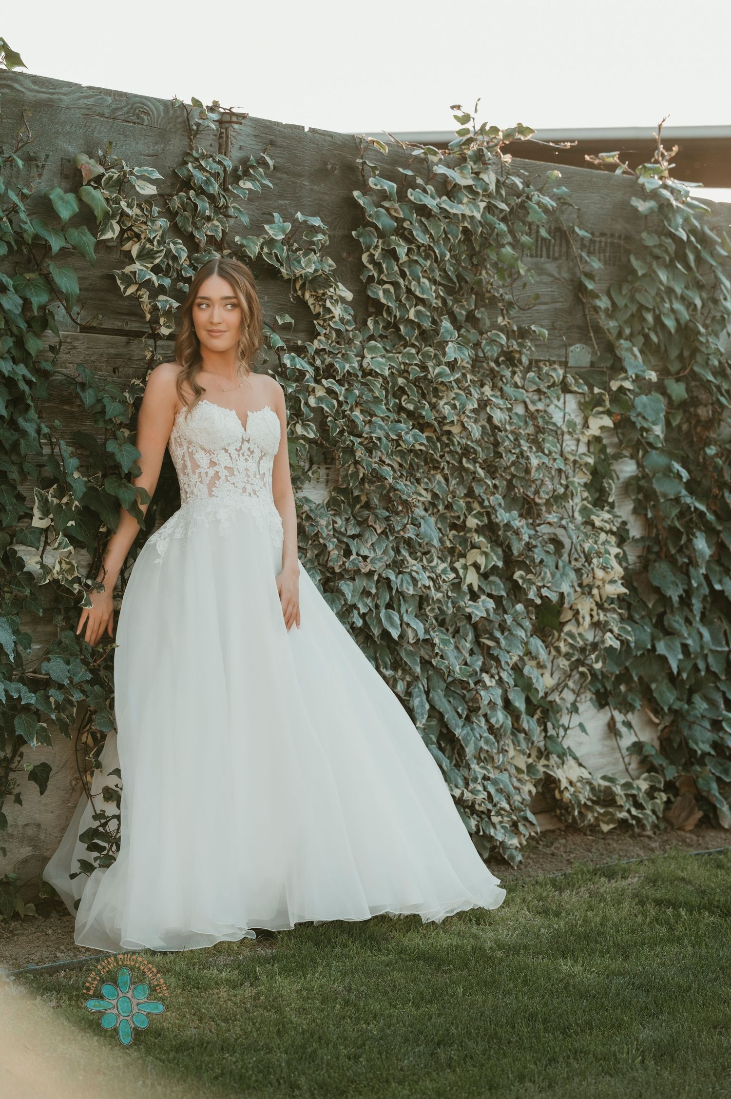 A bride in a flowing white gown poses against an ivy-covered wall at sunset.