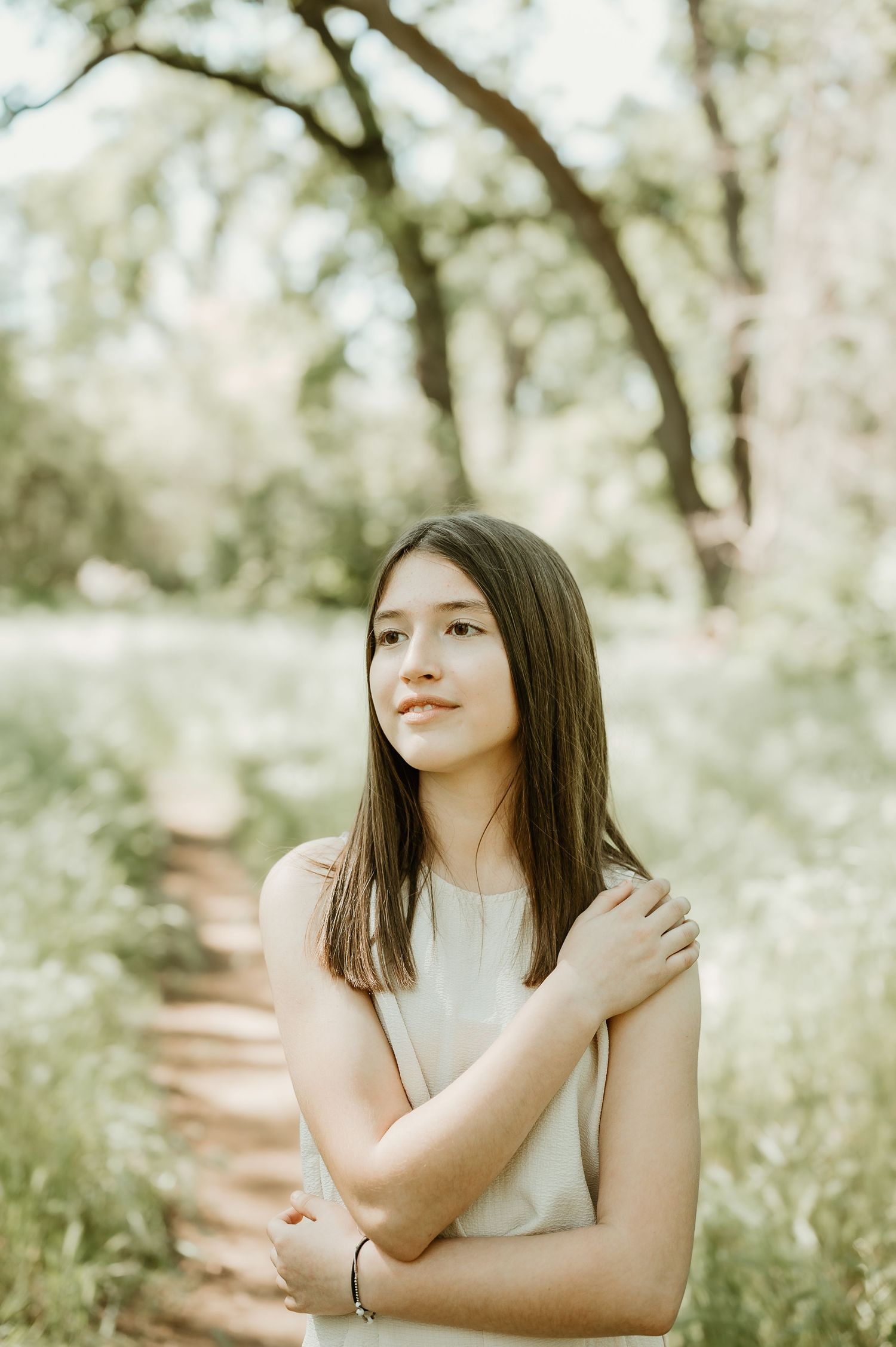 Portrait photograph in natural lighting of someone wearing a sleeveless white top standing on a wooded path surrounded by greenery.