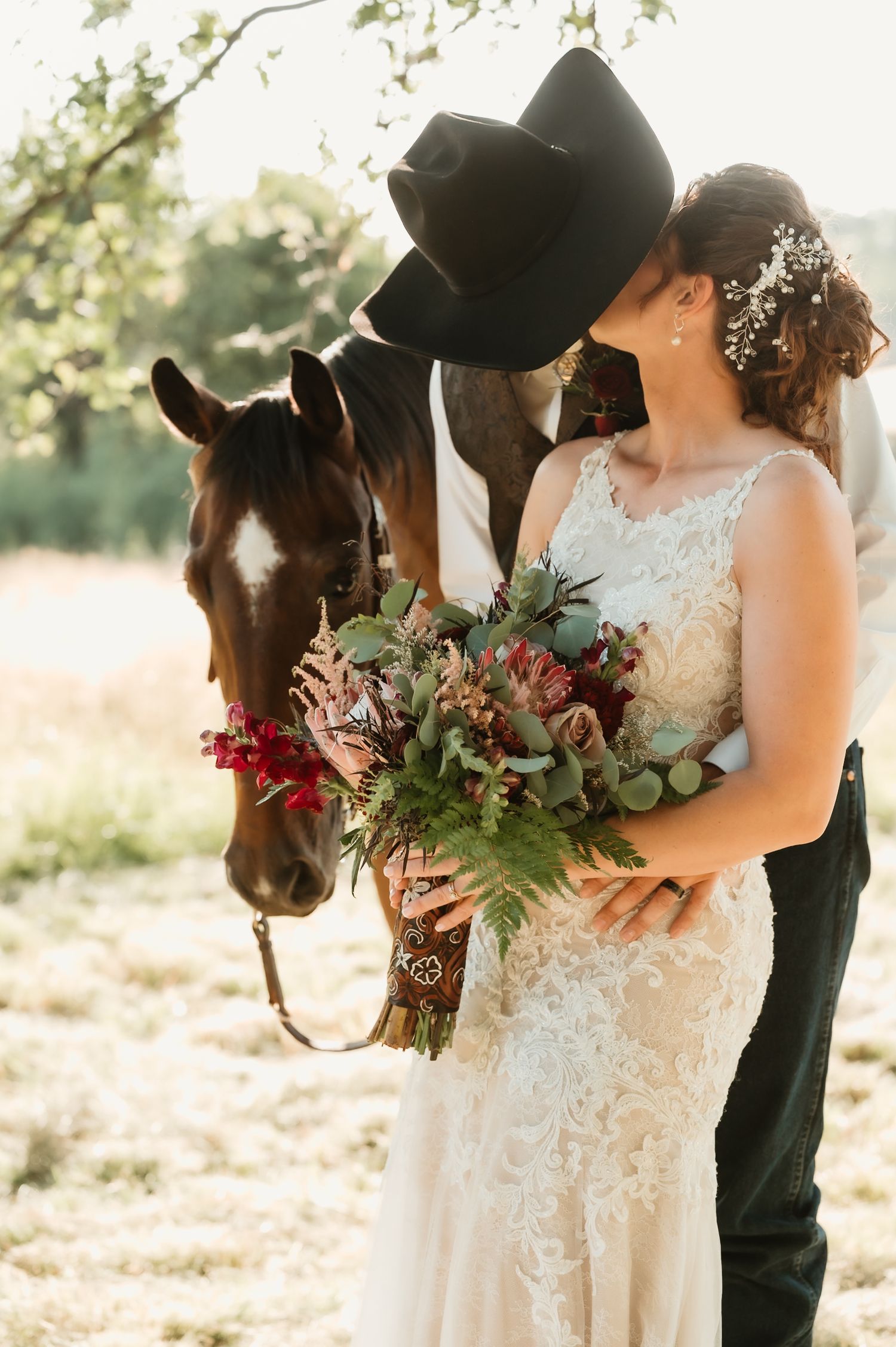 Romantic wedding couple poses with brown horse in rustic outdoor setting at sunset with floral bouquet.