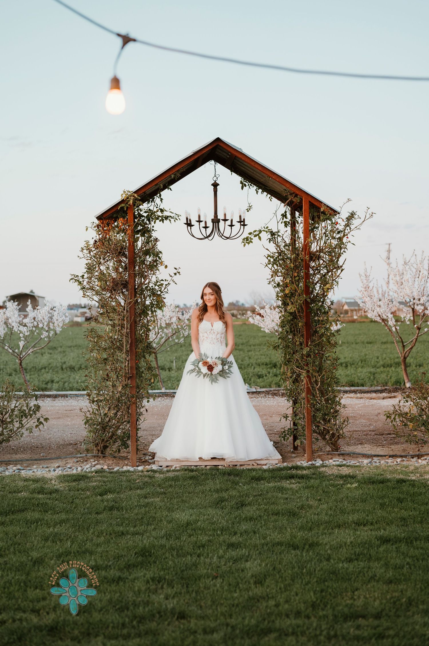 Bride standing under wooden arch decorated with greenery and chandelier in garden setting.