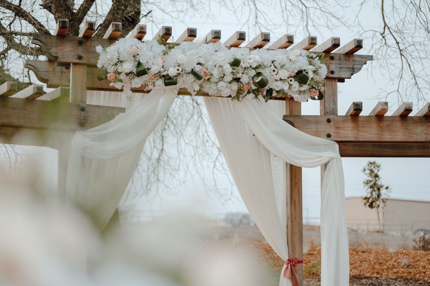 White fabric drapes over a wooden pergola decorated for an outdoor wedding ceremony.