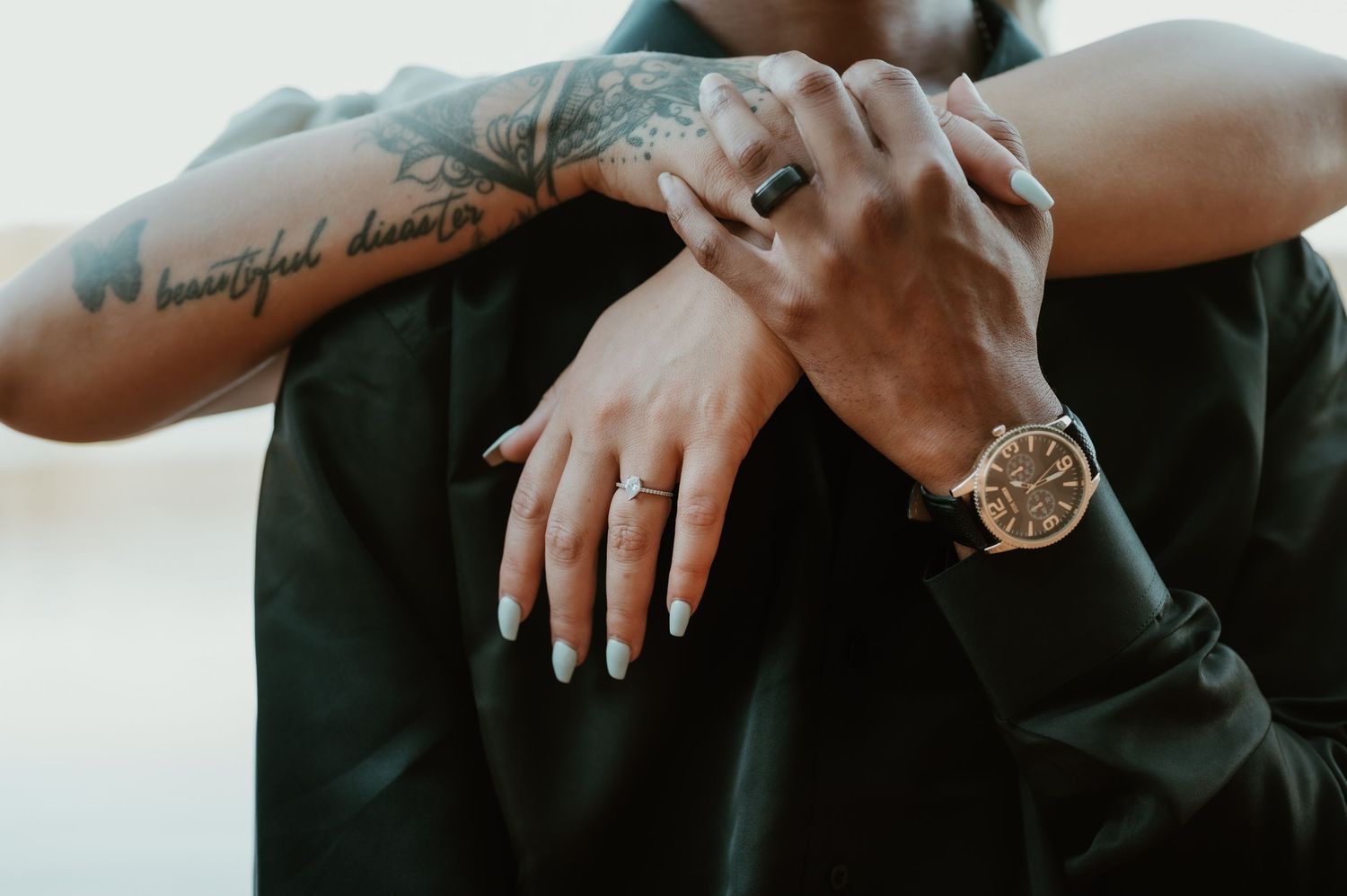 Close-up of hands with visible tattoo and gold watch against dark clothing in soft lighting.