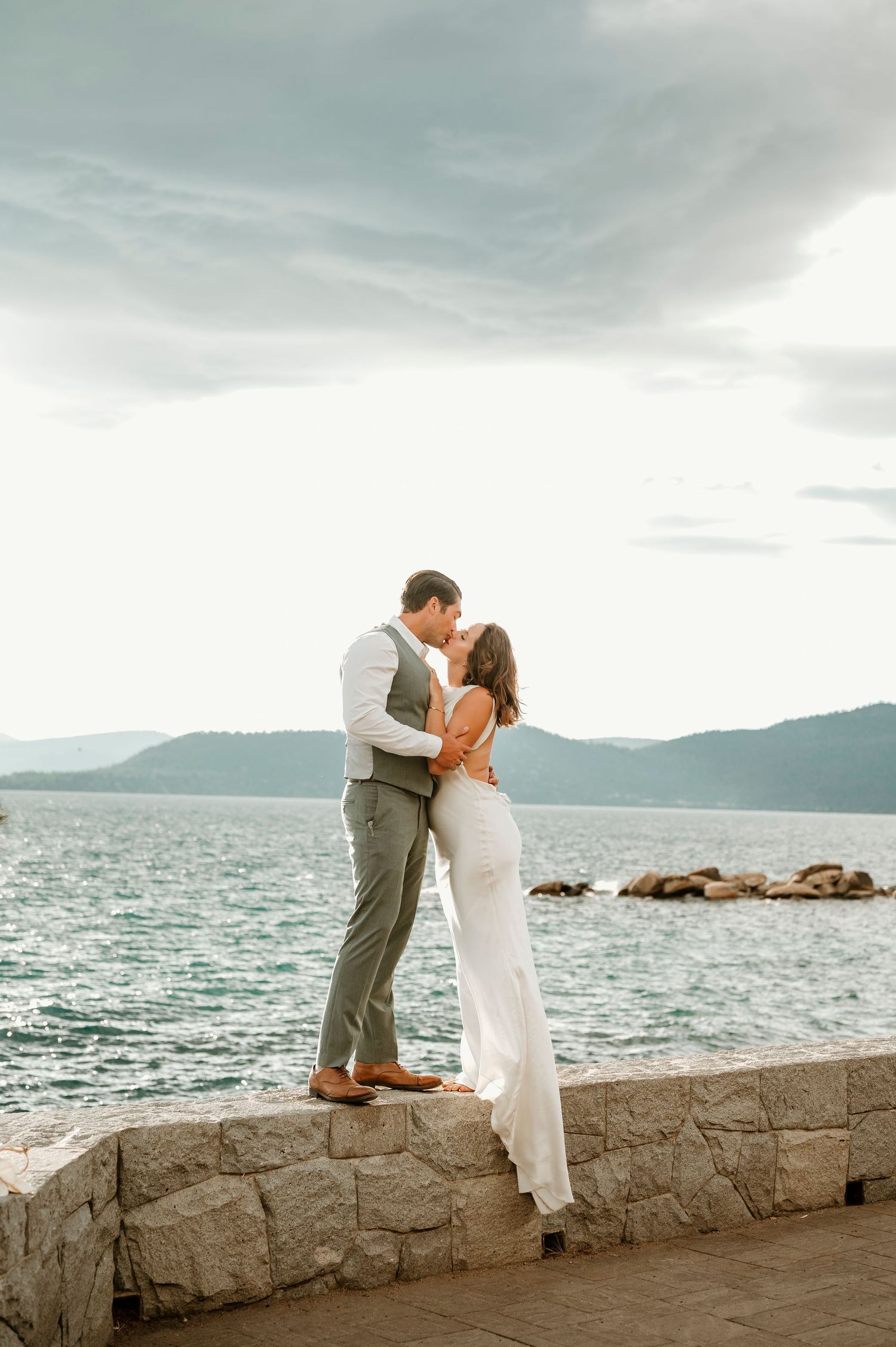 A couple shares an intimate moment on a seaside wall with mountains in the background.