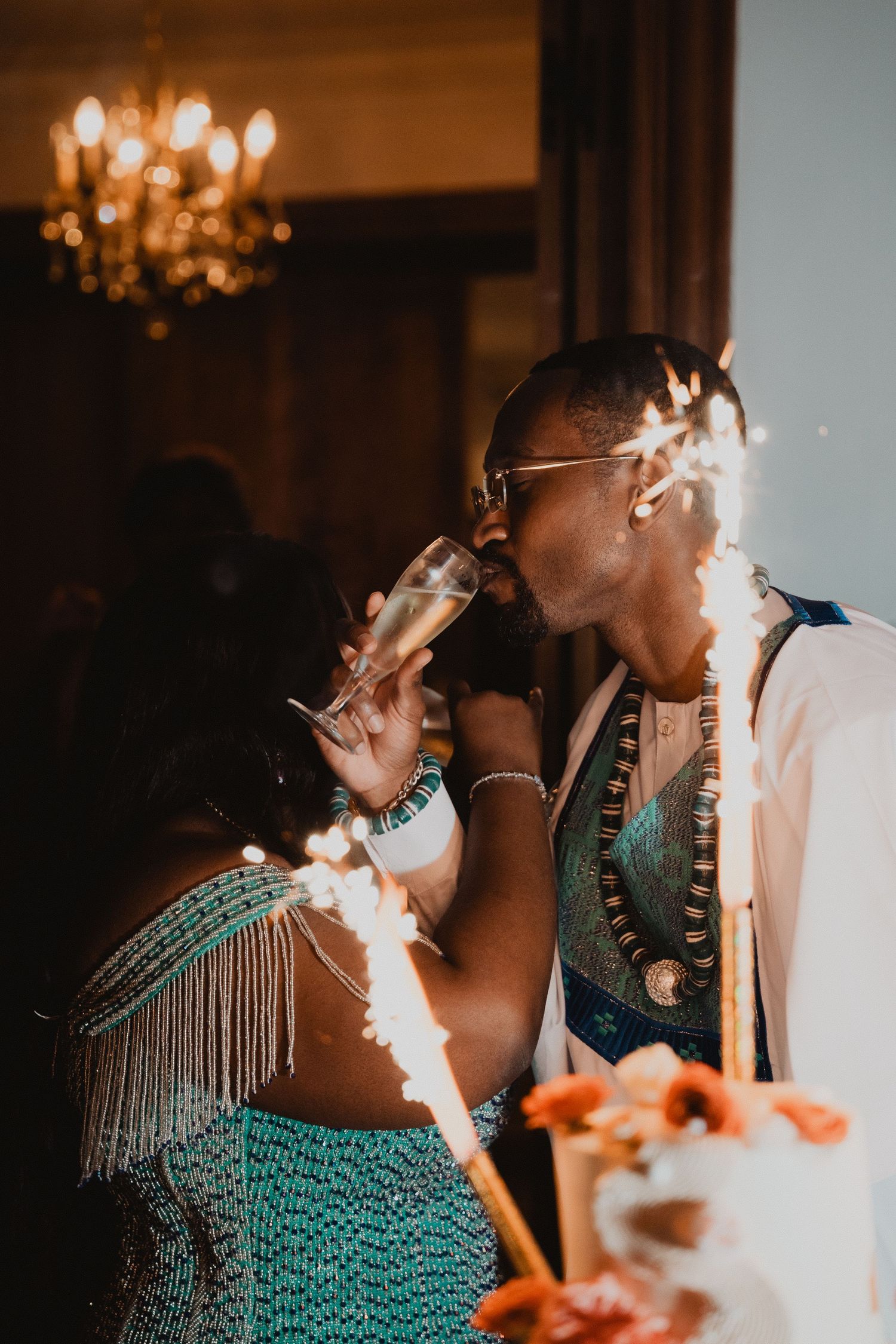 Une personne en robe à franges sirote une boisson lors d'une élégante soirée, avec un lustre en arrière-plan.