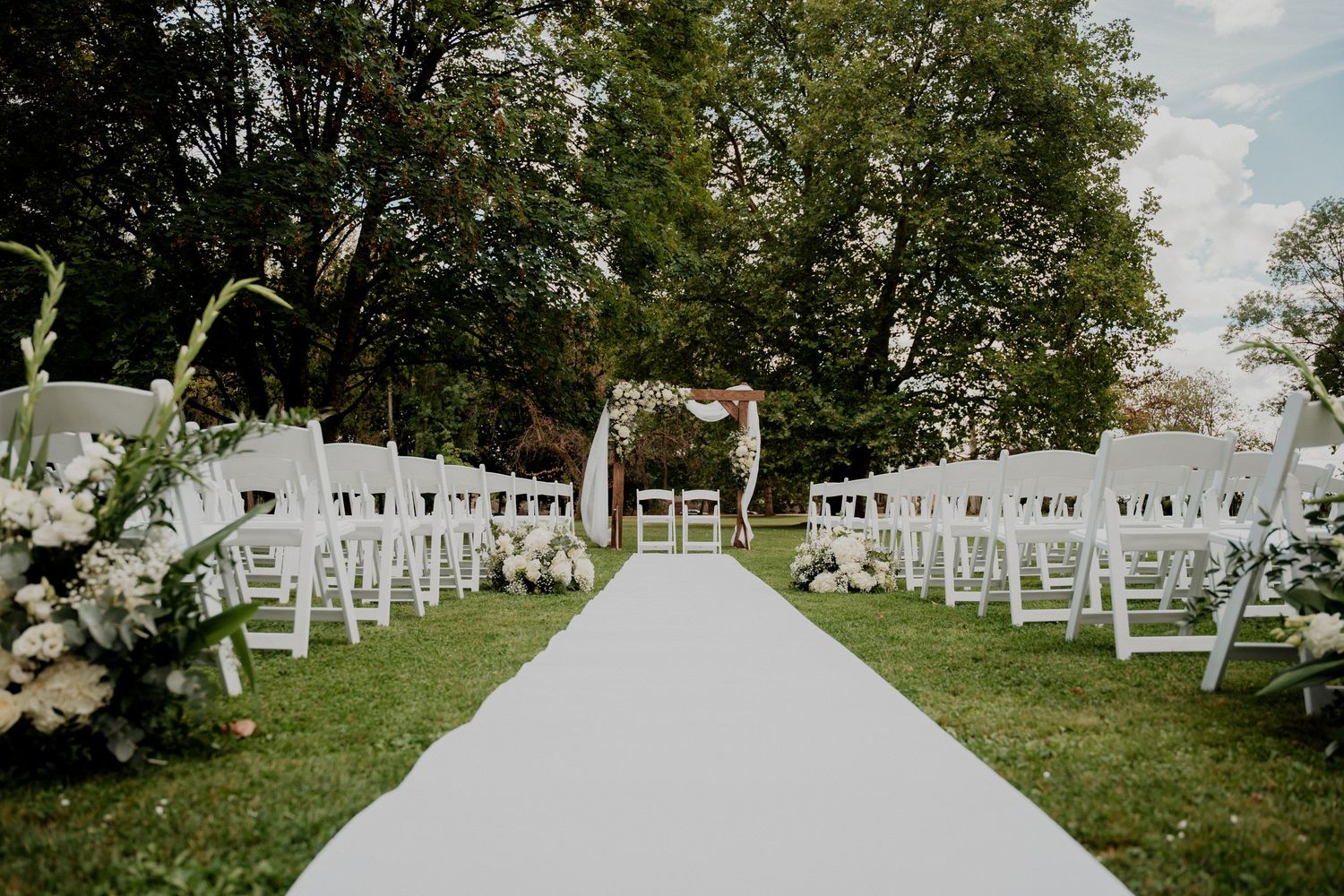 Cérémonie de mariage en plein air, avec des chaises blanches disposées sur une pelouse sous les arbres.