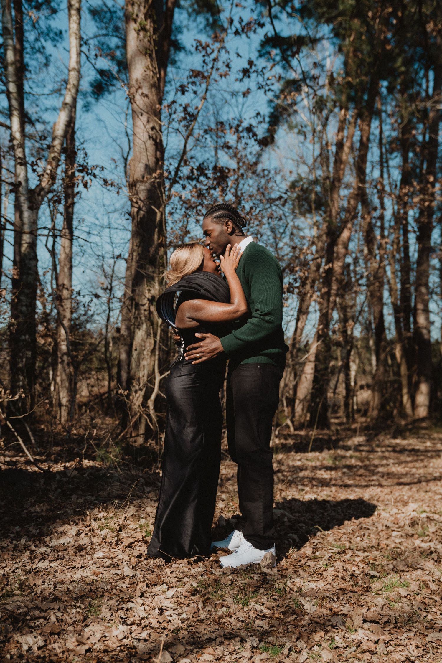 Un couple s'enlace dans une clairière ensoleillée, entourée de grands arbres.
