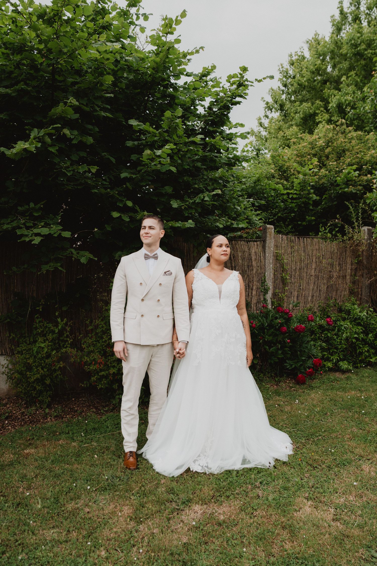 Un couple en tenue de mariage pose dans un jardin avec une clôture rustique en arrière-plan.