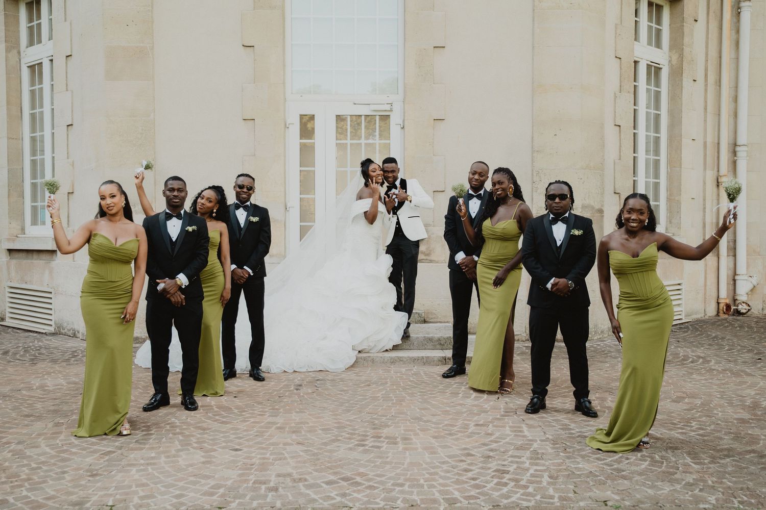 Les membres du cortège nuptial, vêtus de robes vert olive et de costumes noirs, posent le long du mur blanc du bâtiment.