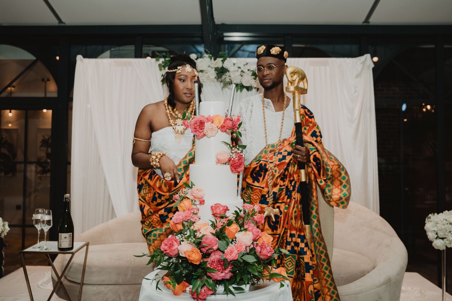 Un couple coupe son gâteau de mariage décoré de fleurs éclatantes, vêtu de tenues traditionnelles africaines.