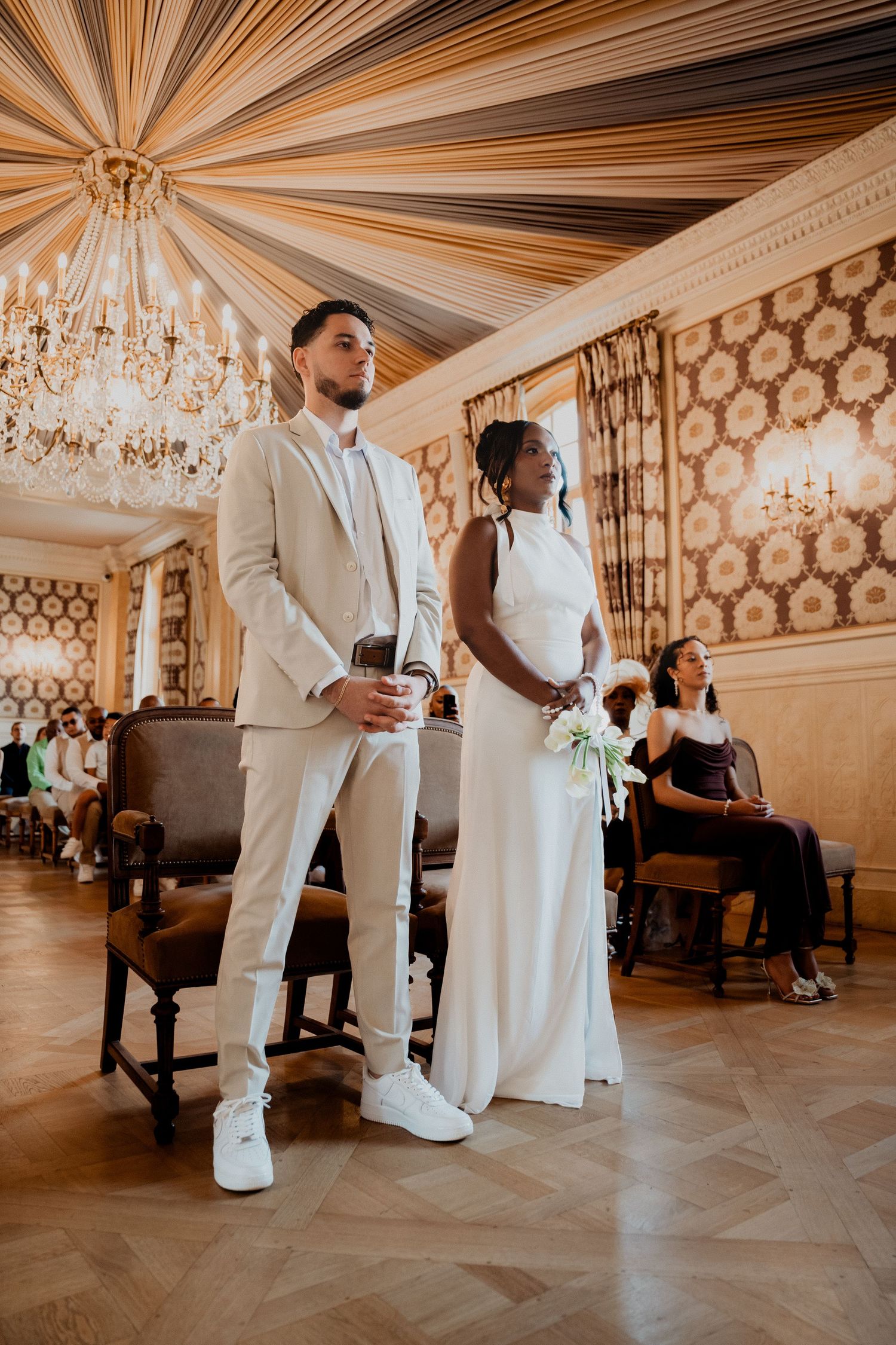 Un couple se tient debout dans une élégante salle de bal au plafond drapé et aux lustres en cristal pendant leur cérémonie.