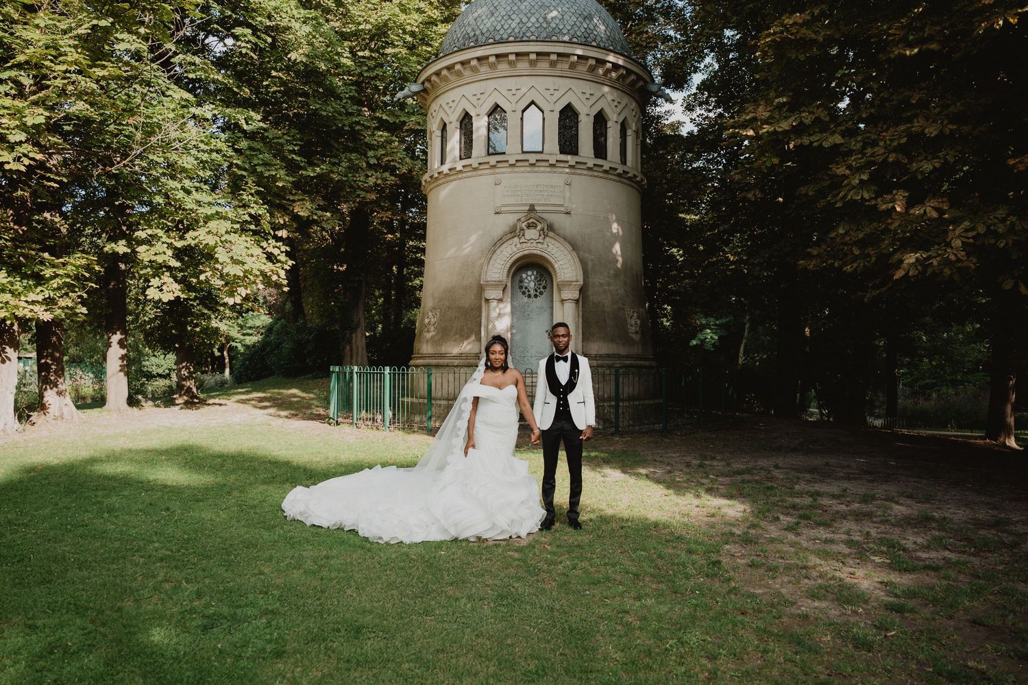 A wedding couple poses in front of a historic stone tower on a sunlit lawn.