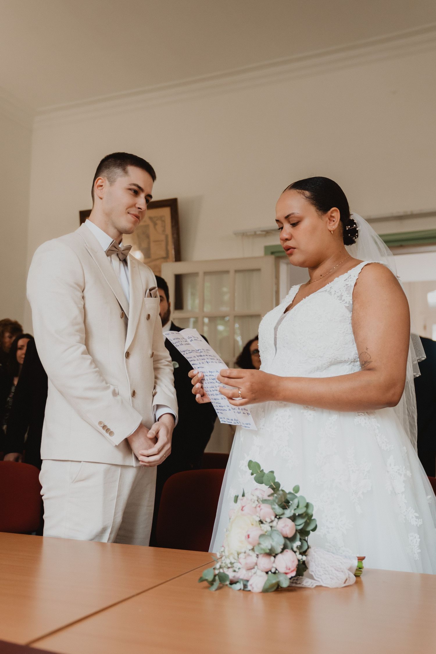 Une cérémonie de mariage est en cours ; un couple échange ses vœux debout autour d'une table décorée de fleurs.