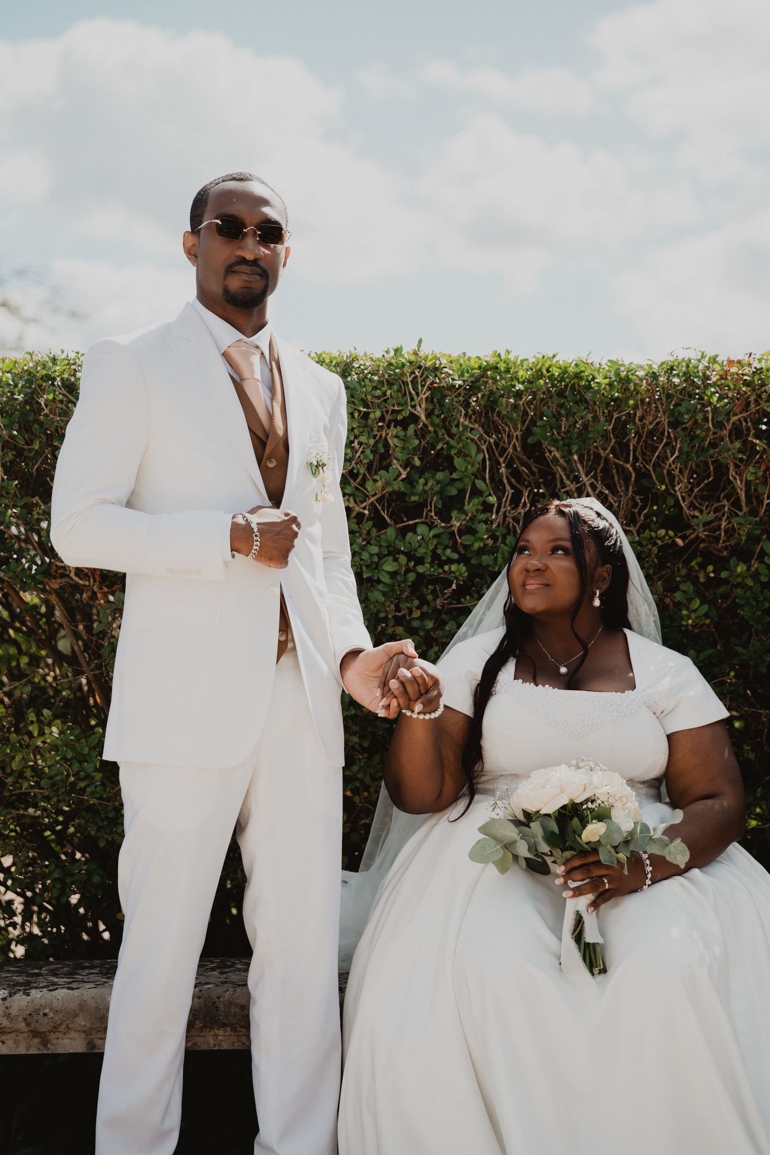 Wedding couple in elegant white attire posing against a green hedge backdrop.