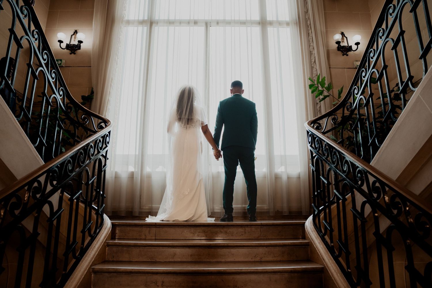 Un couple se détache en silhouette en haut d'un élégant escalier aux rampes ornées, avec une grande fenêtre en arrière-plan.