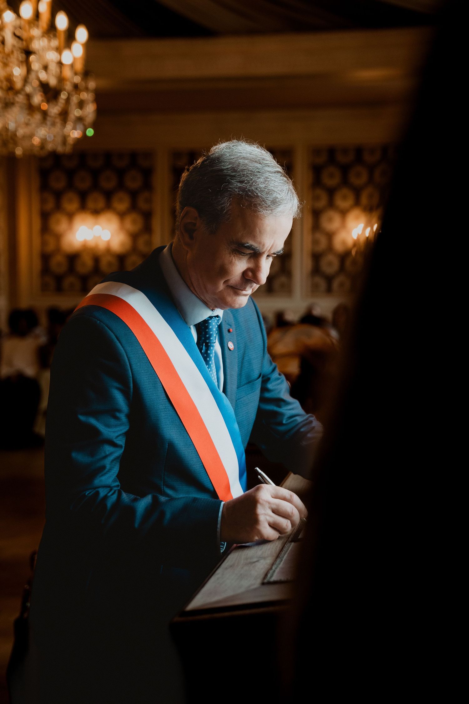 Un maire en costume bleu et écharpe tricolore signe des documents dans une pièce richement décorée de lustres.