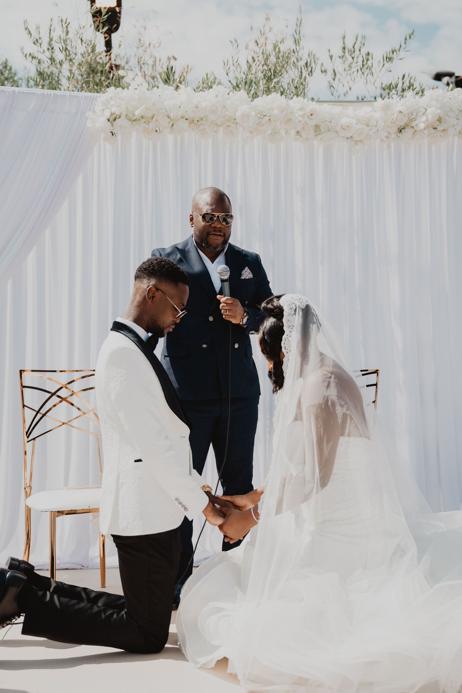 Un moment intime de cérémonie de mariage avec le marié agenouillé et l'officiant devant un drapé blanc.