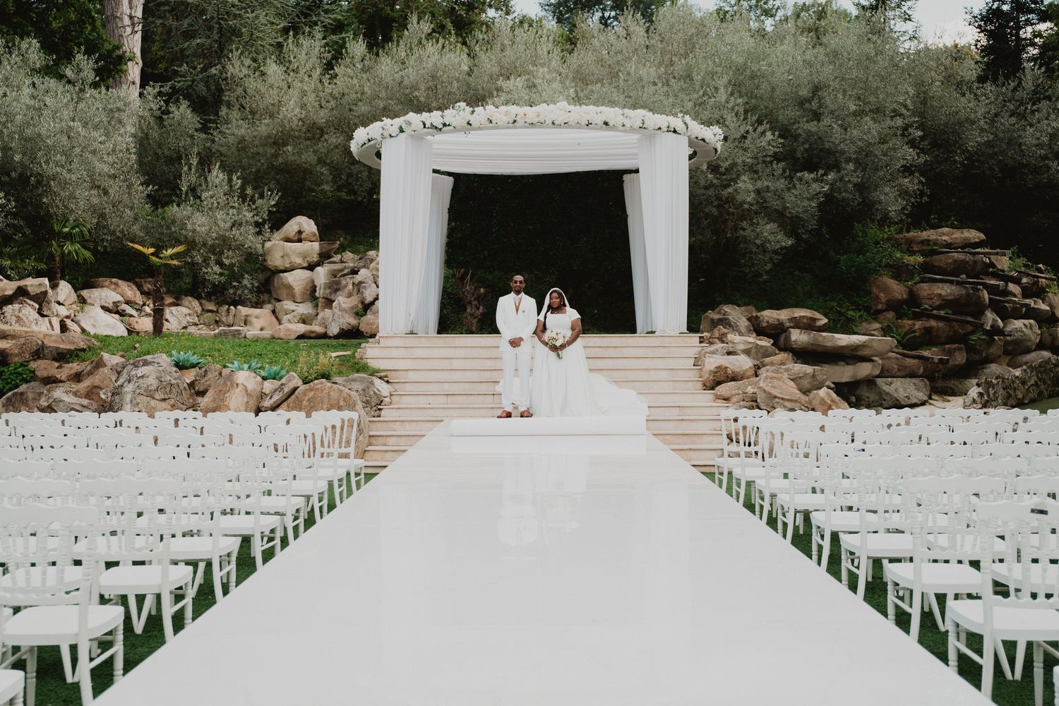 Elegant outdoor wedding ceremony setup with white chairs and floral arrangements at a garden venue.