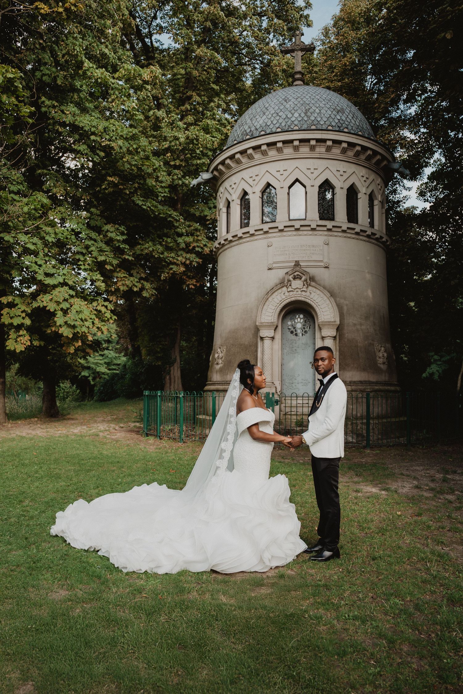 Newlyweds hold hands in front of an ornate stone tower surrounded by lush greenery.