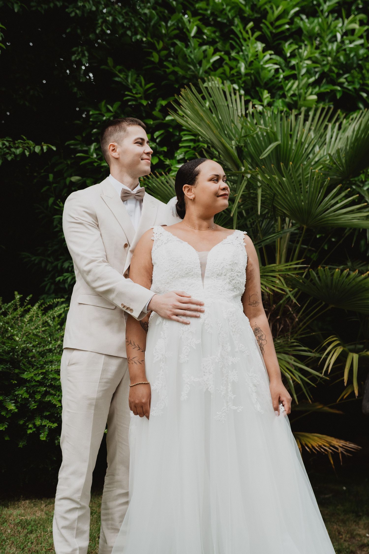 Wedding couple in white attire poses together in a tropical garden setting.