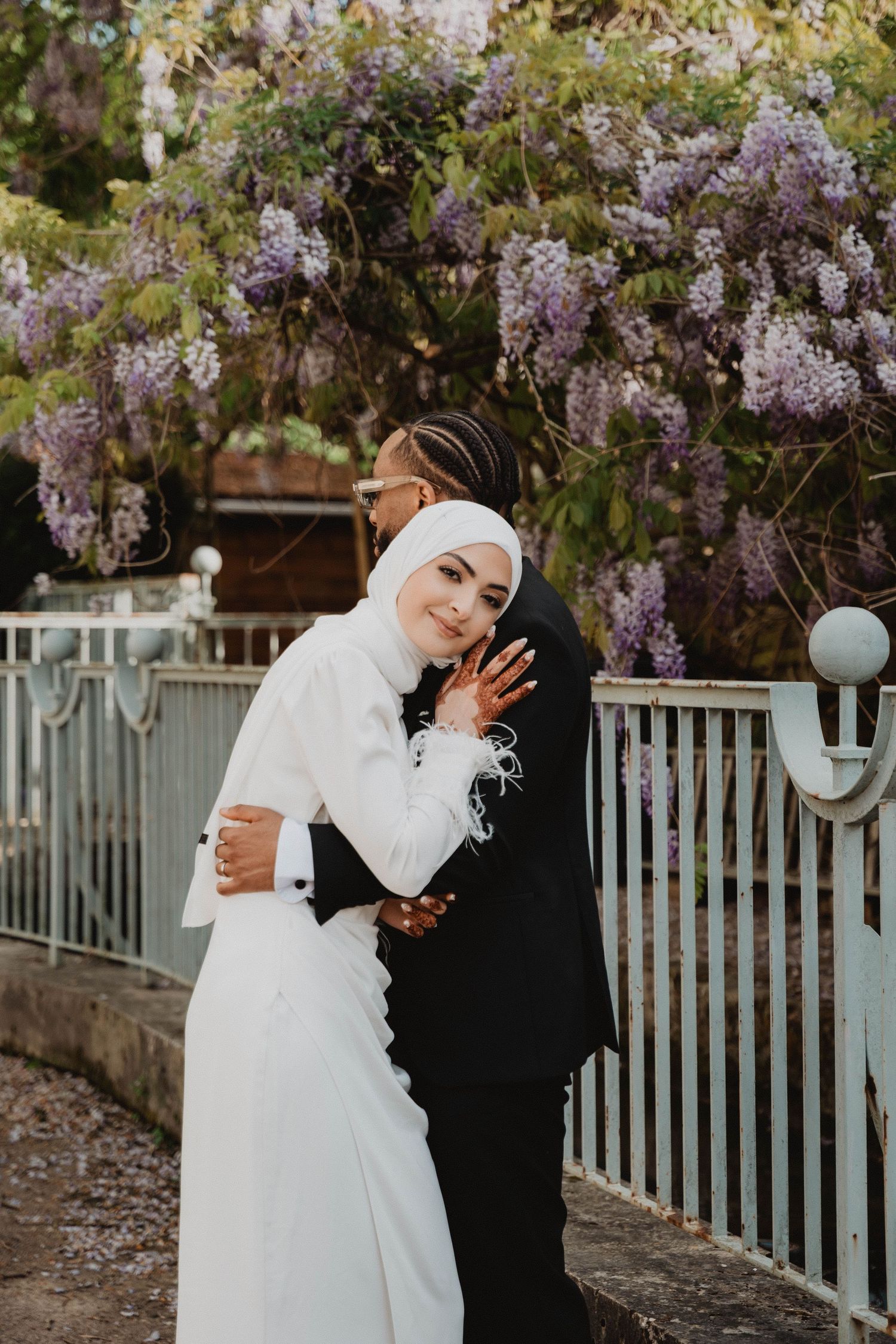 Un couple s'enlace sous une glycine en fleurs et une clôture blanche.