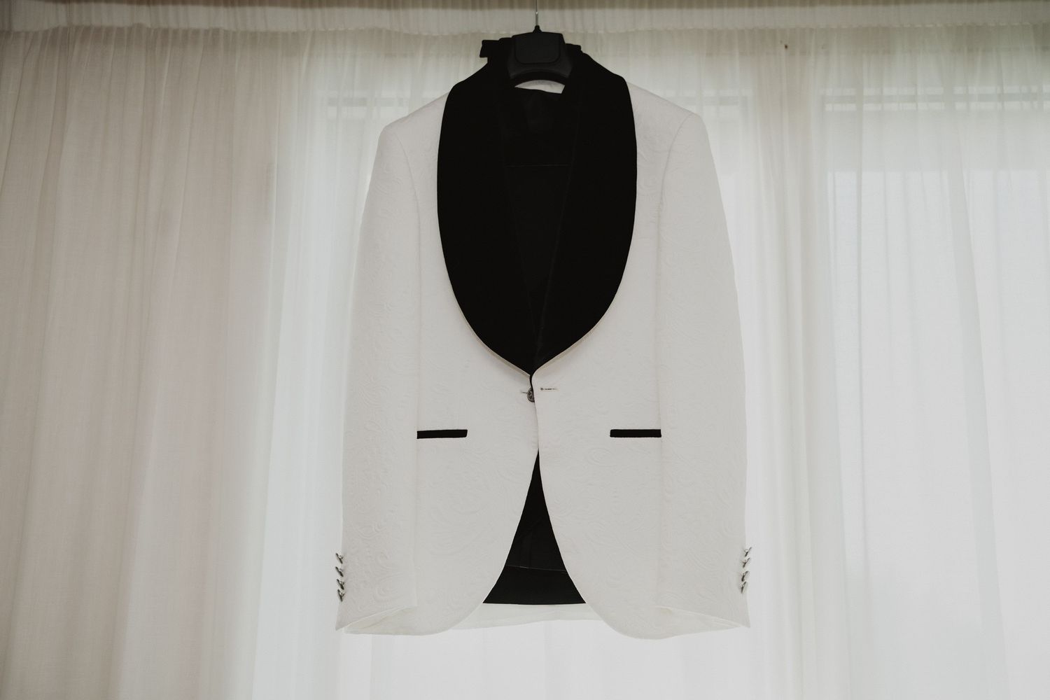 Une veste de smoking blanche à revers noirs, suspendue à un cintre devant un rideau blanc.