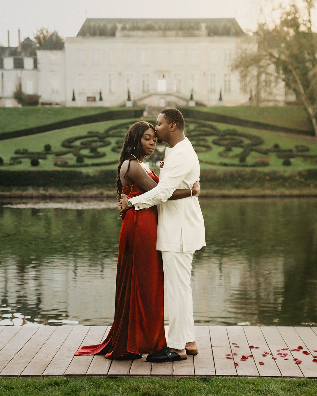 Un couple en tenue de soirée s'embrasse sur le quai, avec un élégant bâtiment blanc et des jardins soignés en arrière-plan.