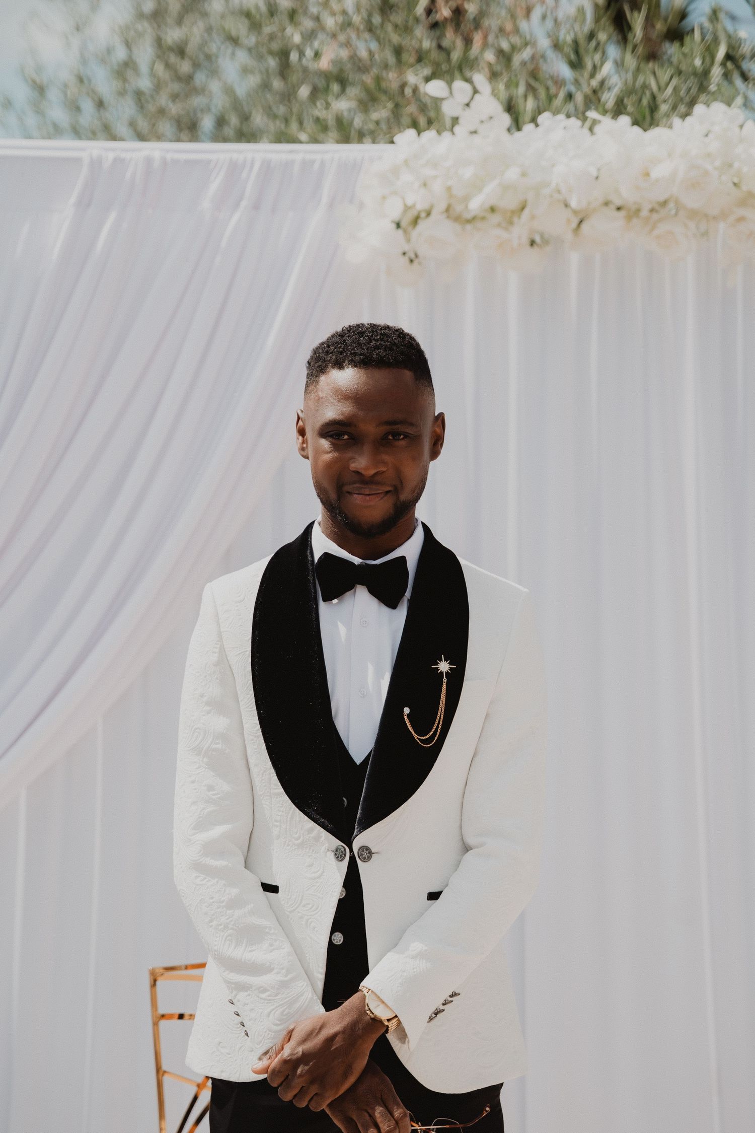 Un marié en smoking blanc à revers noirs sourit lors d'une cérémonie de mariage en plein air.