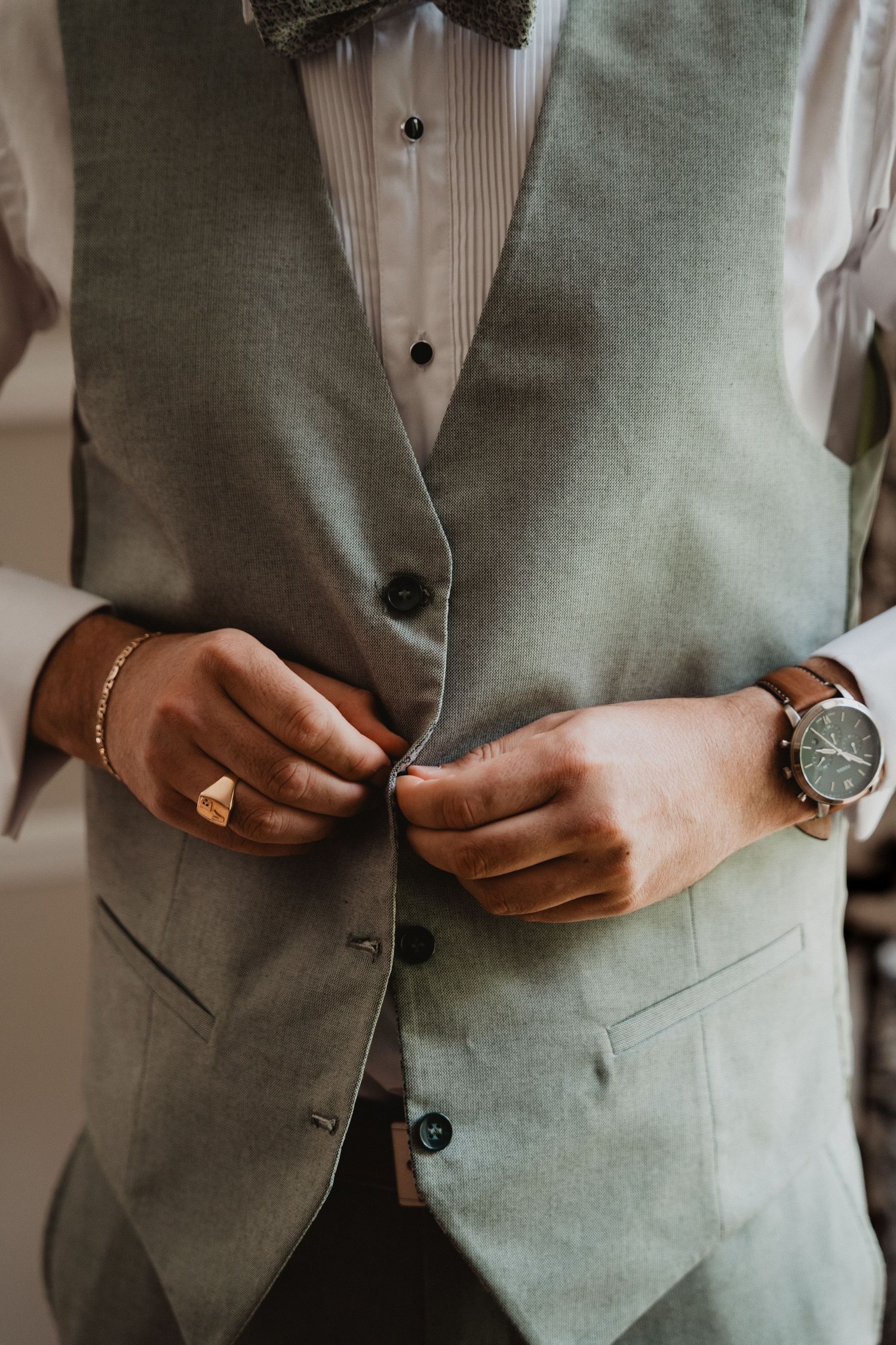 Gros plan sur le torse d'un marié montrant un gilet gris, une montre et ses mains boutonnant sa chemise.